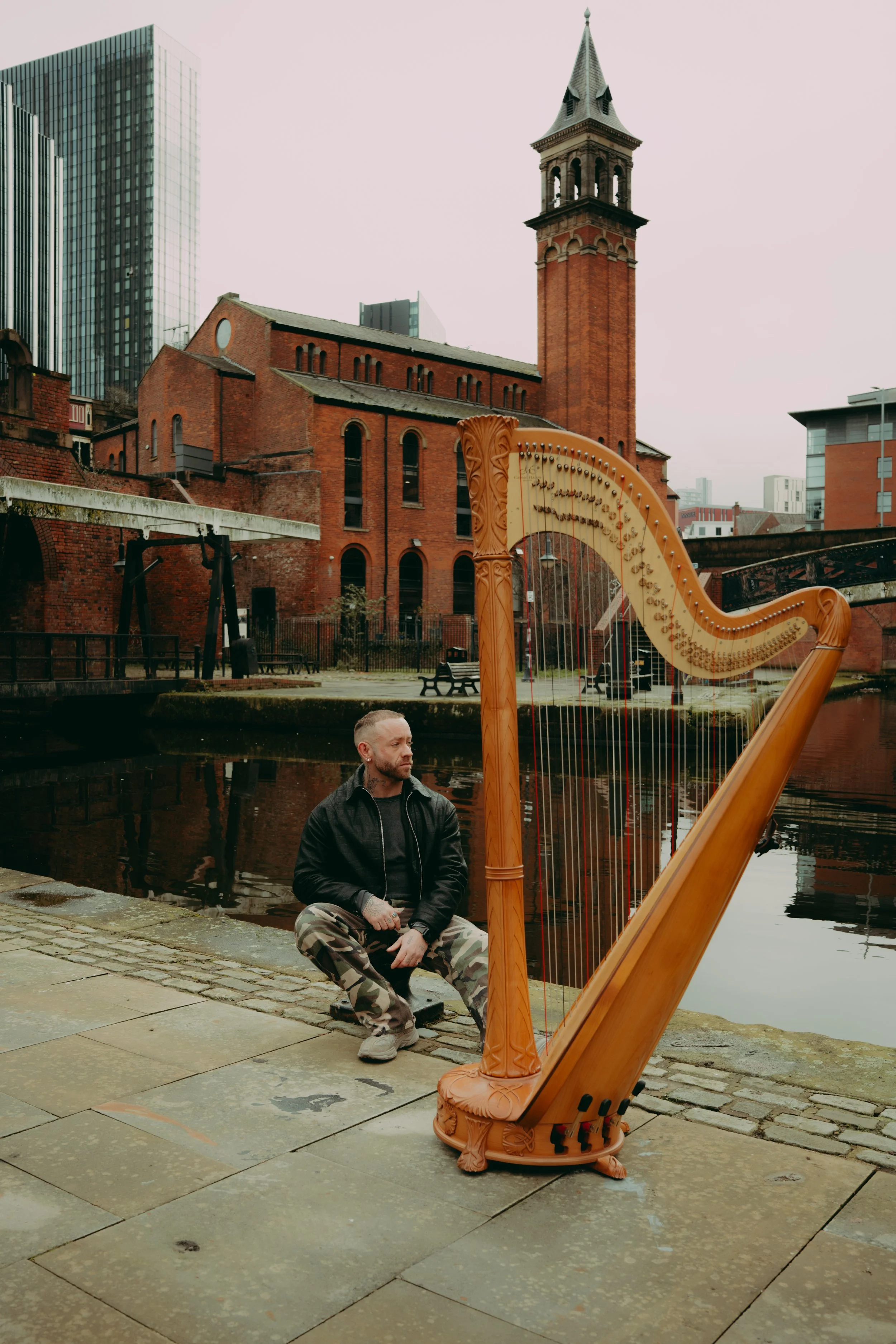 A man with a beard in a black leather jacket and camouflage pants sits on the cobblestone sidewalk beside a large  harp, near a body of water with reflections of buildings, including an old brick church with a tower, and modern skyscrapers in the background. The sky is overcast.
