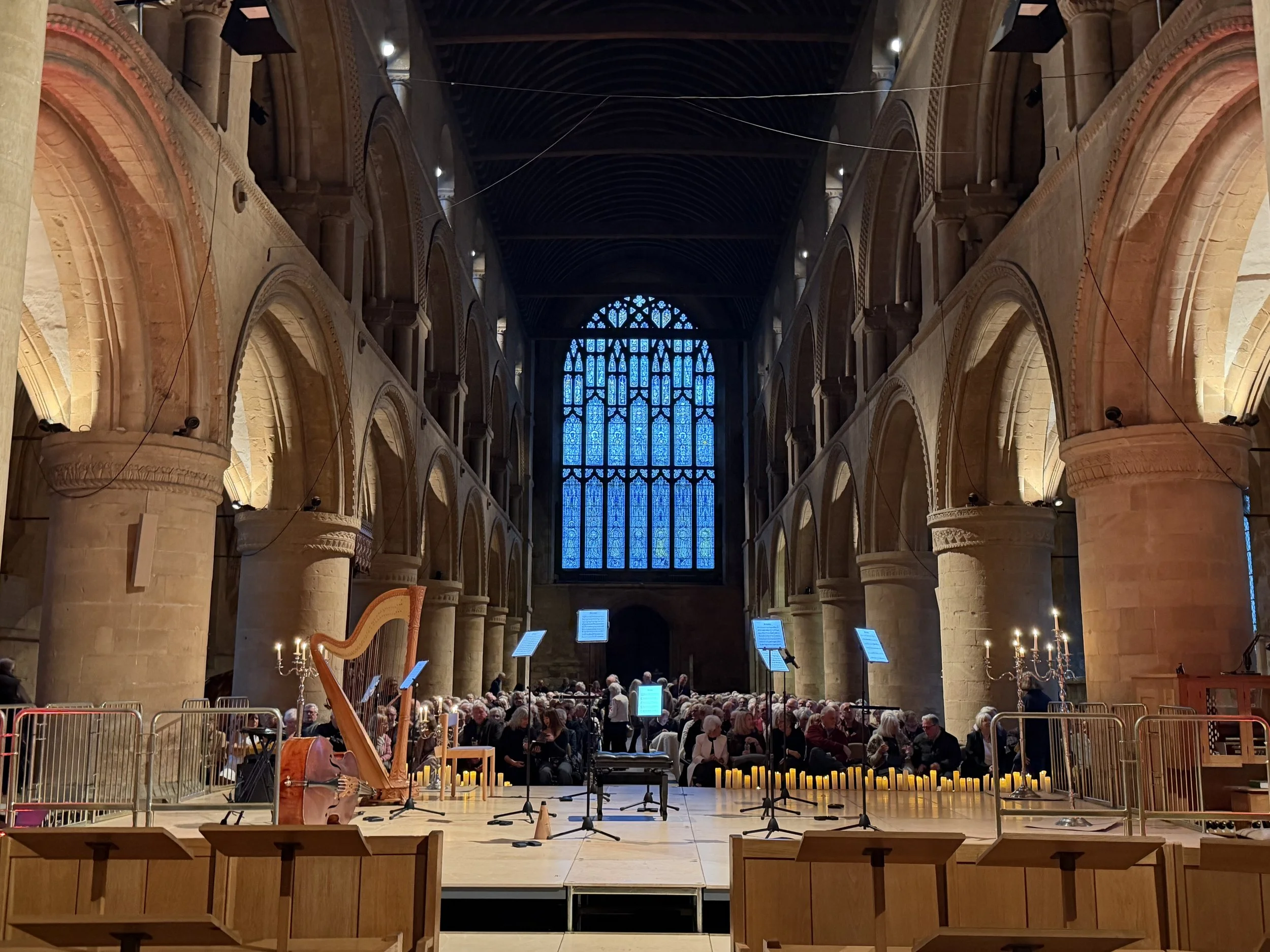 Inside a church or cathedral, a large congregation seated for a performance or service, with musical stands and instruments including a harp and microphone stands at the front, stained glass windows high above, and candles lining the stage area.