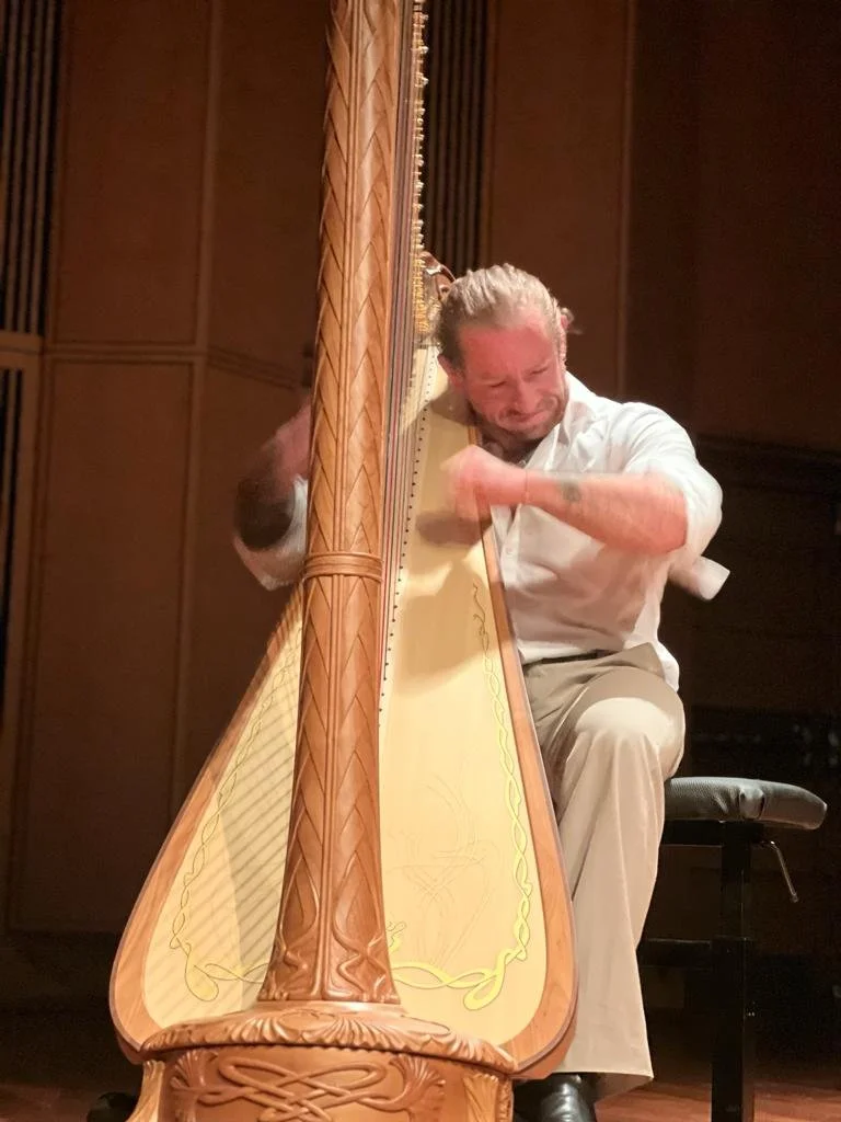 Man playing a harp on stage, seated on a chair, with a focused or emotional expression.