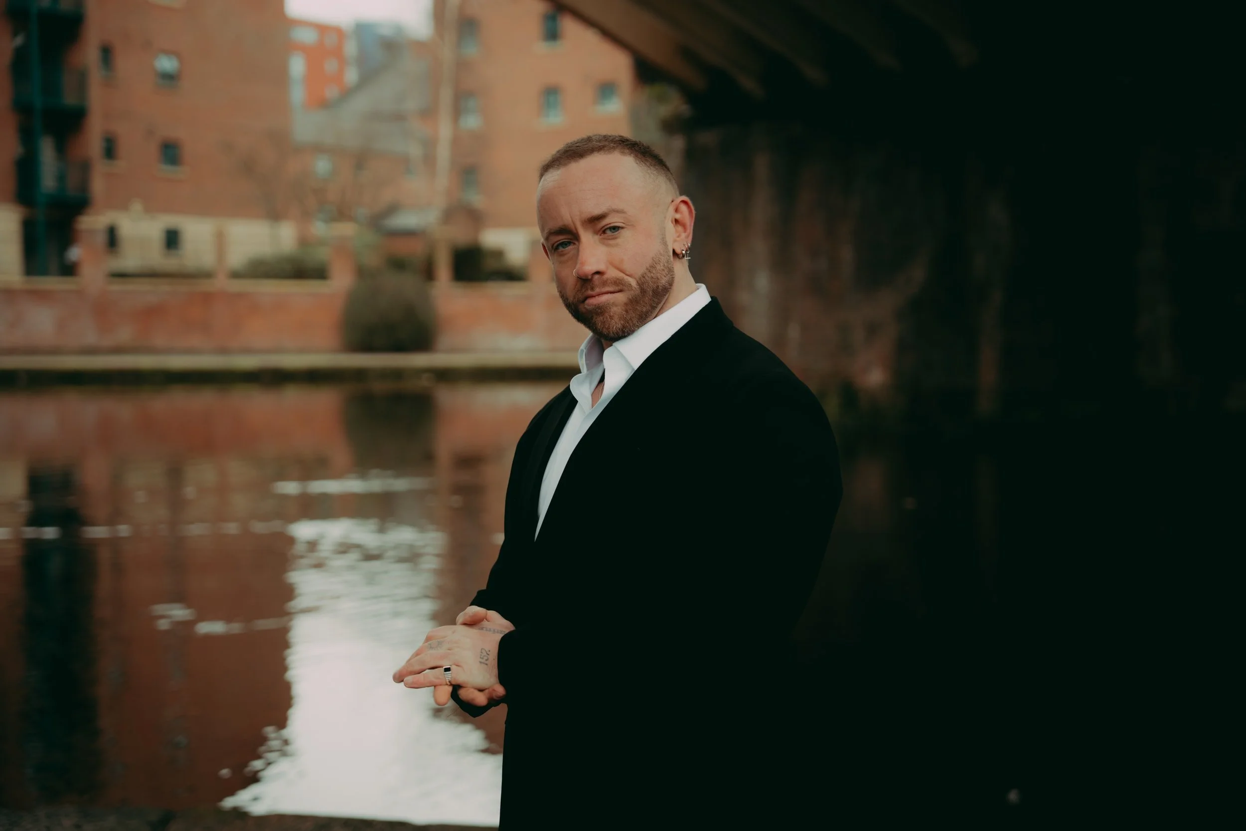 A man with a short beard and earrings wearing a black suit and white shirt, standing outdoors near a body of water with brick buildings and a bridge in the background.