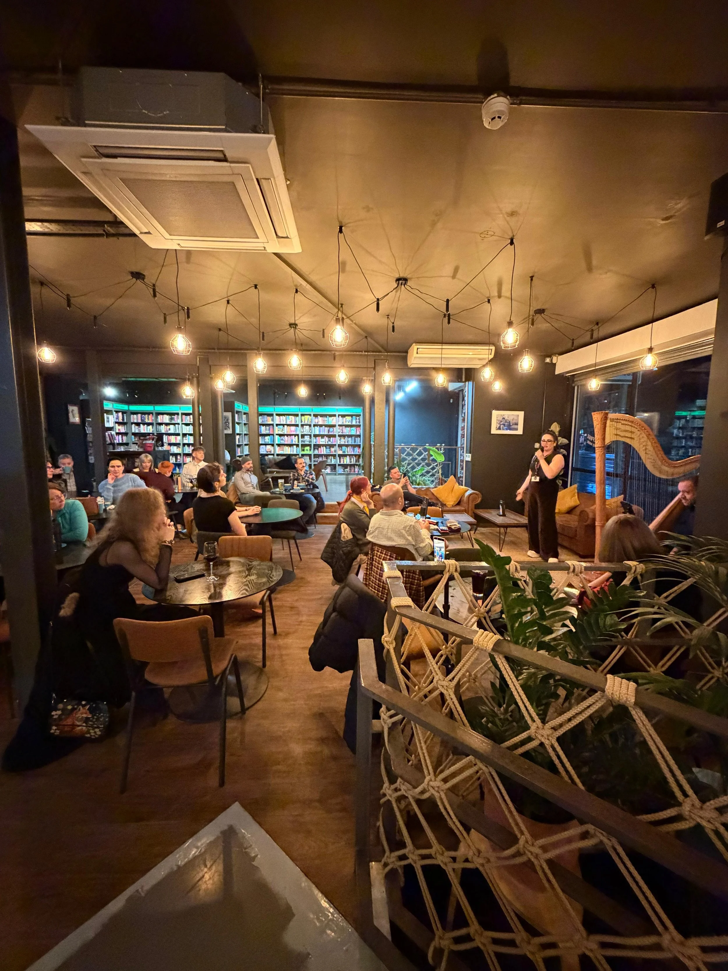 Indoor gathering in a cozy, dimly lit space with people sitting at tables and a woman speaking into a microphone, with bookshelves and warm lighting.