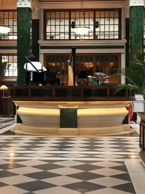 Empty hotel lobby reception desk with musical instruments behind it, including a grand piano and drum set, in a building with large windows and checkered floor.