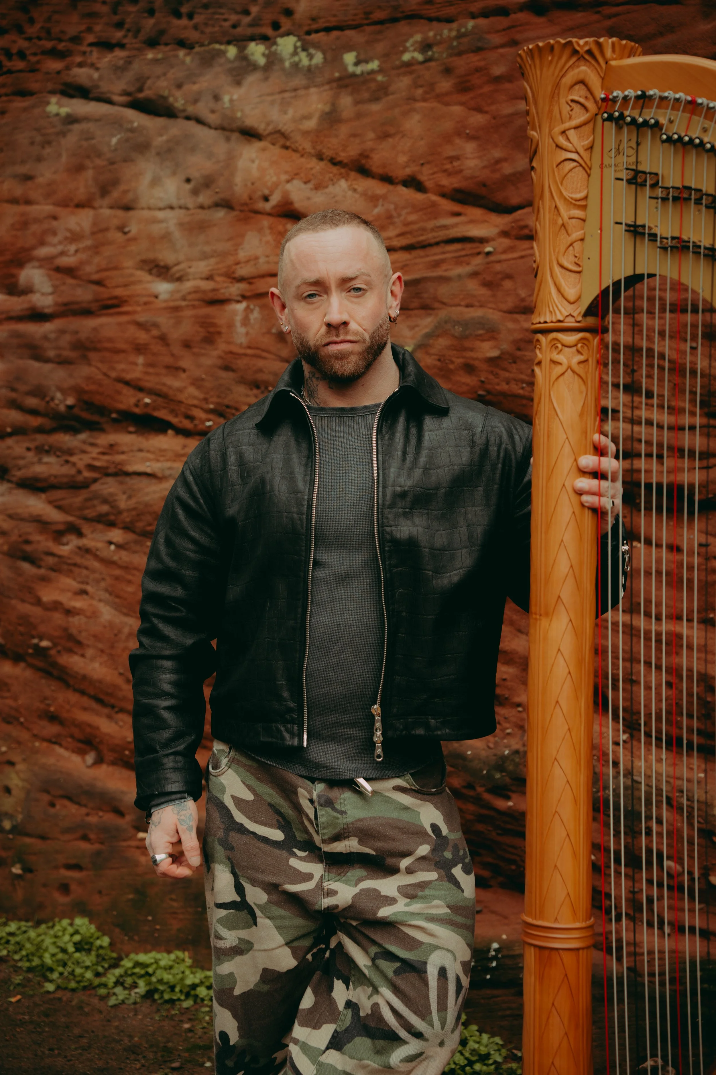 Man with a beard and short hair holding a large wooden harp, standing in front of a red rock wall, wearing a black leather jacket, gray shirt, and camouflage pants.