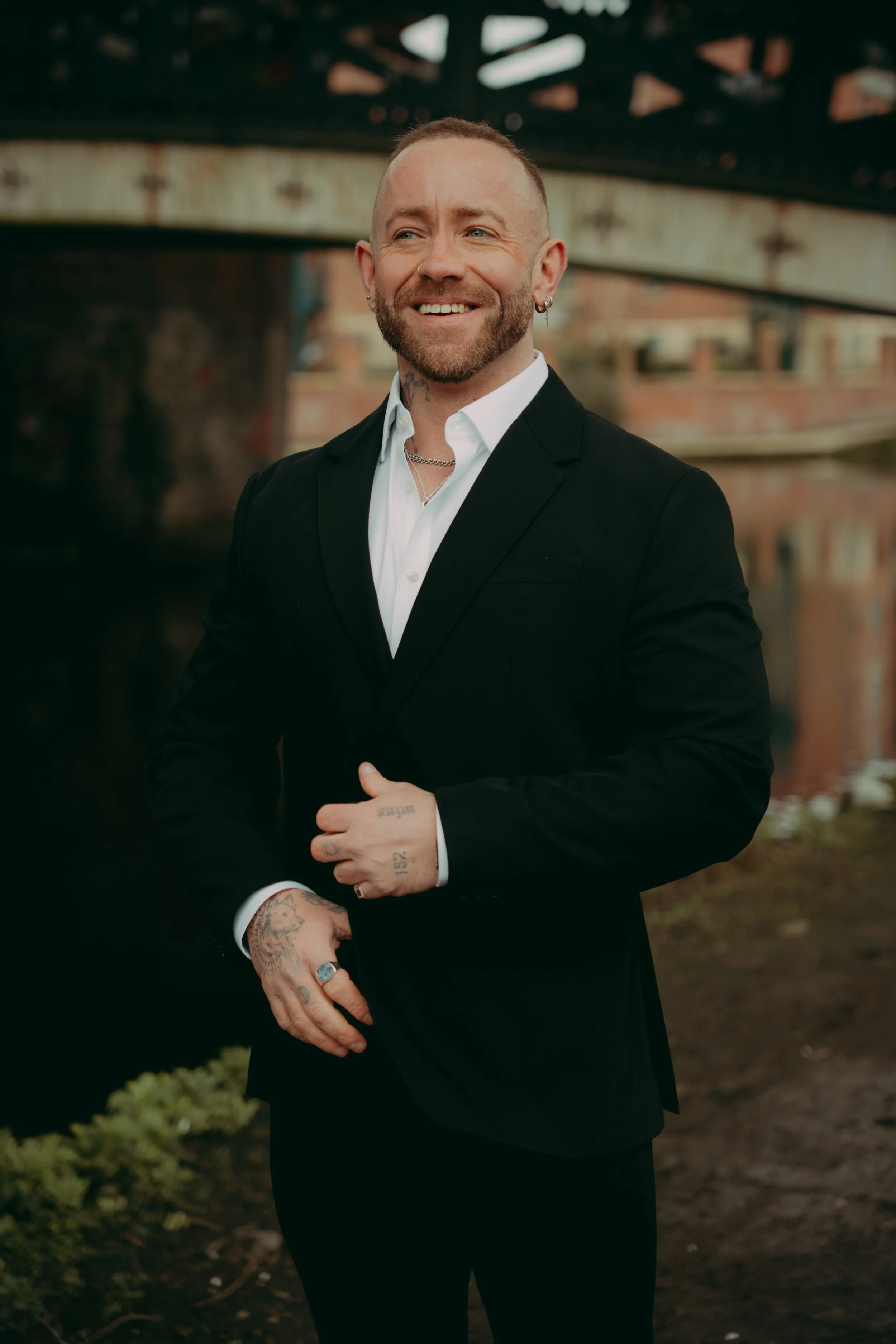 A man with tattoos and earrings dressed in a black suit jacket and white shirt outdoors, smiling with a bridge and water in the background.