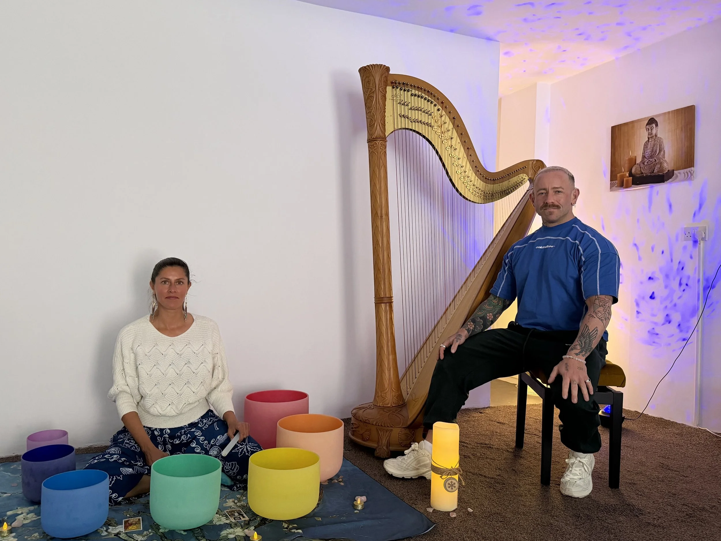 A woman sitting on the floor surrounded by colorful singing bowls and tarot cards, a man sitting on a chair behind a harp, illuminated by soft purple and yellow lights.