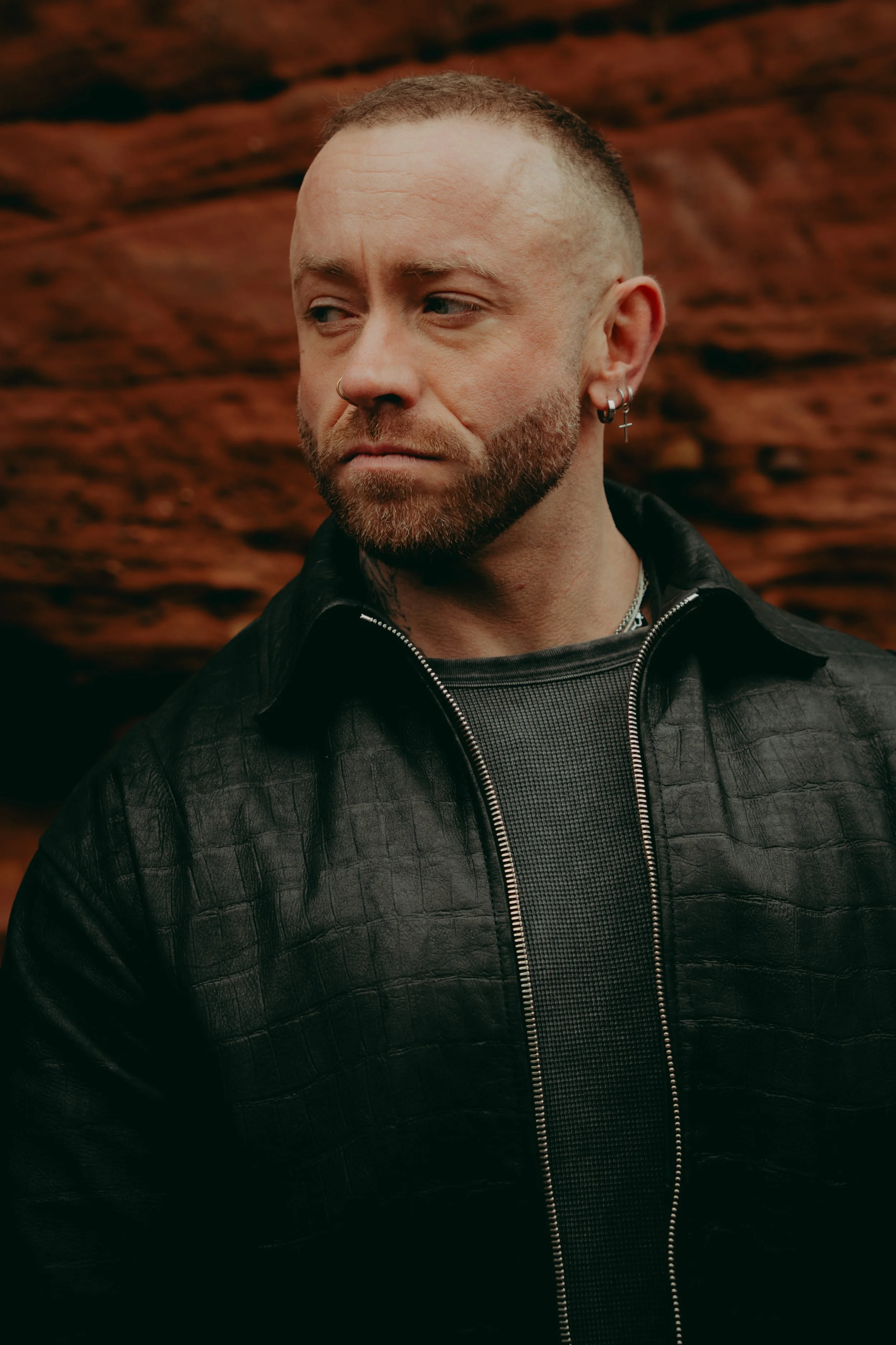 Close-up of a man with short hair and a beard looking to the side, wearing earrings and a black leather jacket, standing in front of a reddish-brown textured background.
