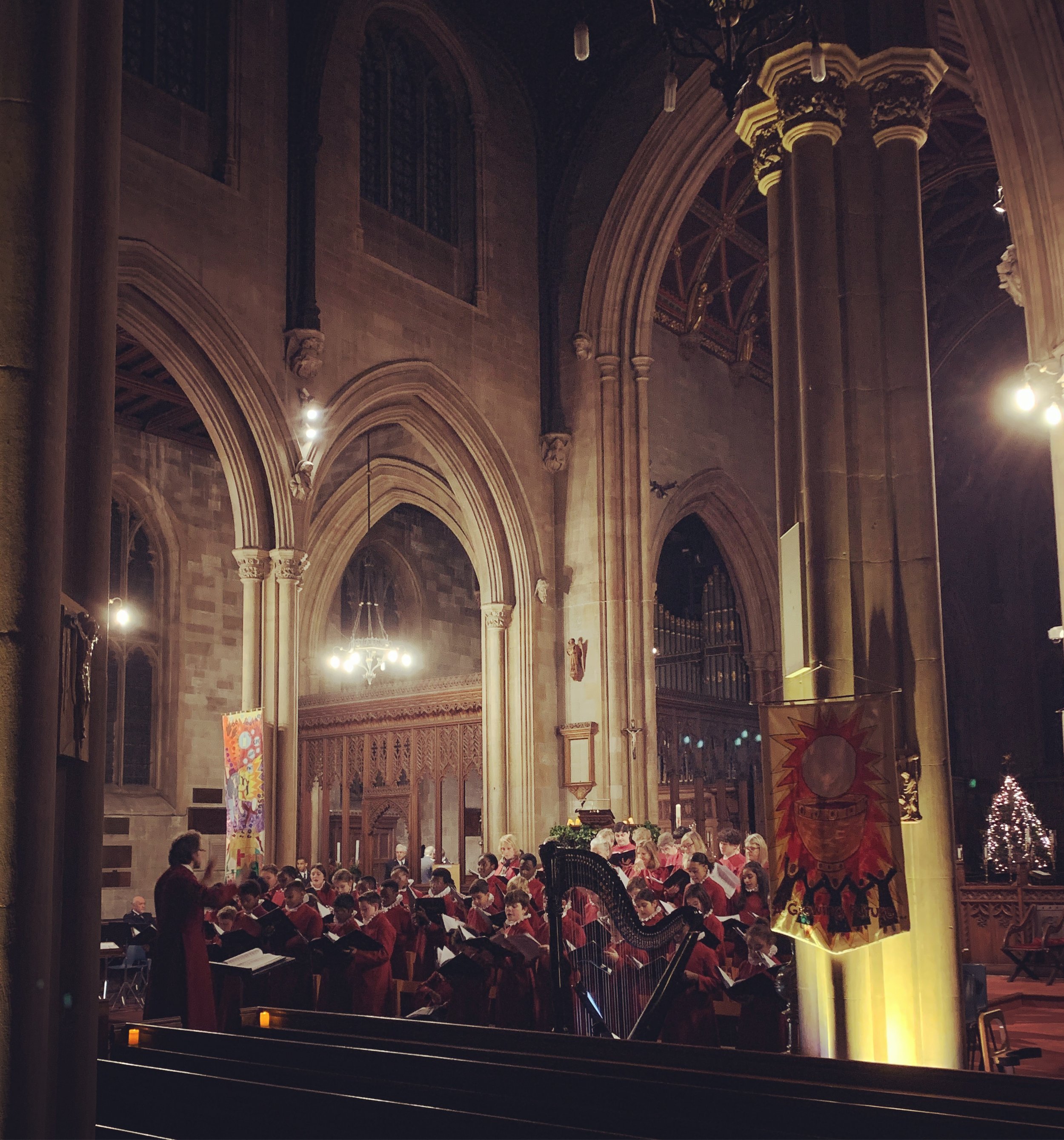 A choir of children dressed in red robes performing in a large, Gothic-style church with high arched ceilings, stained glass windows, and ornate columns. A soloist stands at the front, and a woman conducts the choir. The church is decorated with banners and flags.