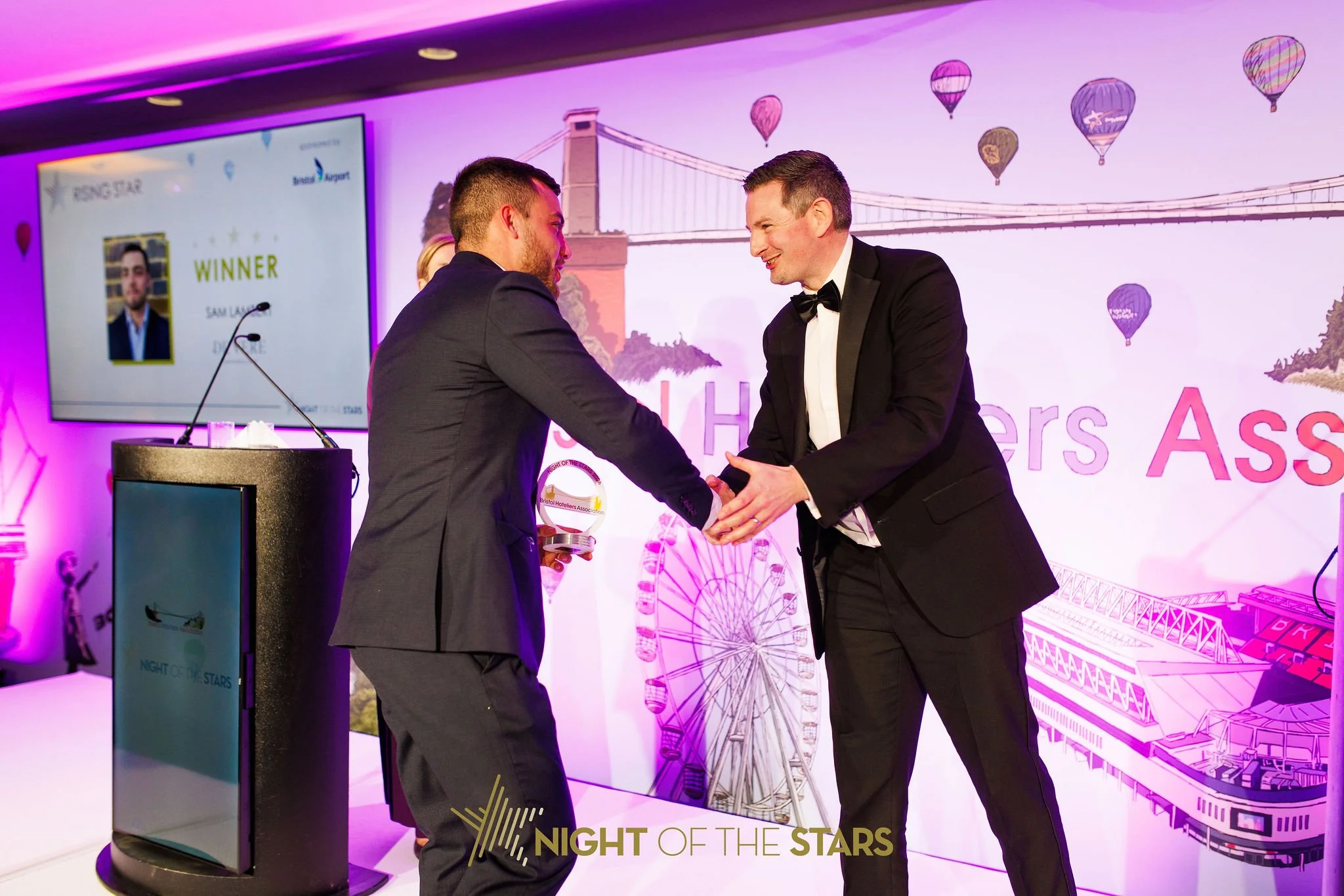Two men in tuxedos shaking hands at an awards event with a colorful carnival backdrop including a Ferris wheel and hot air balloons.