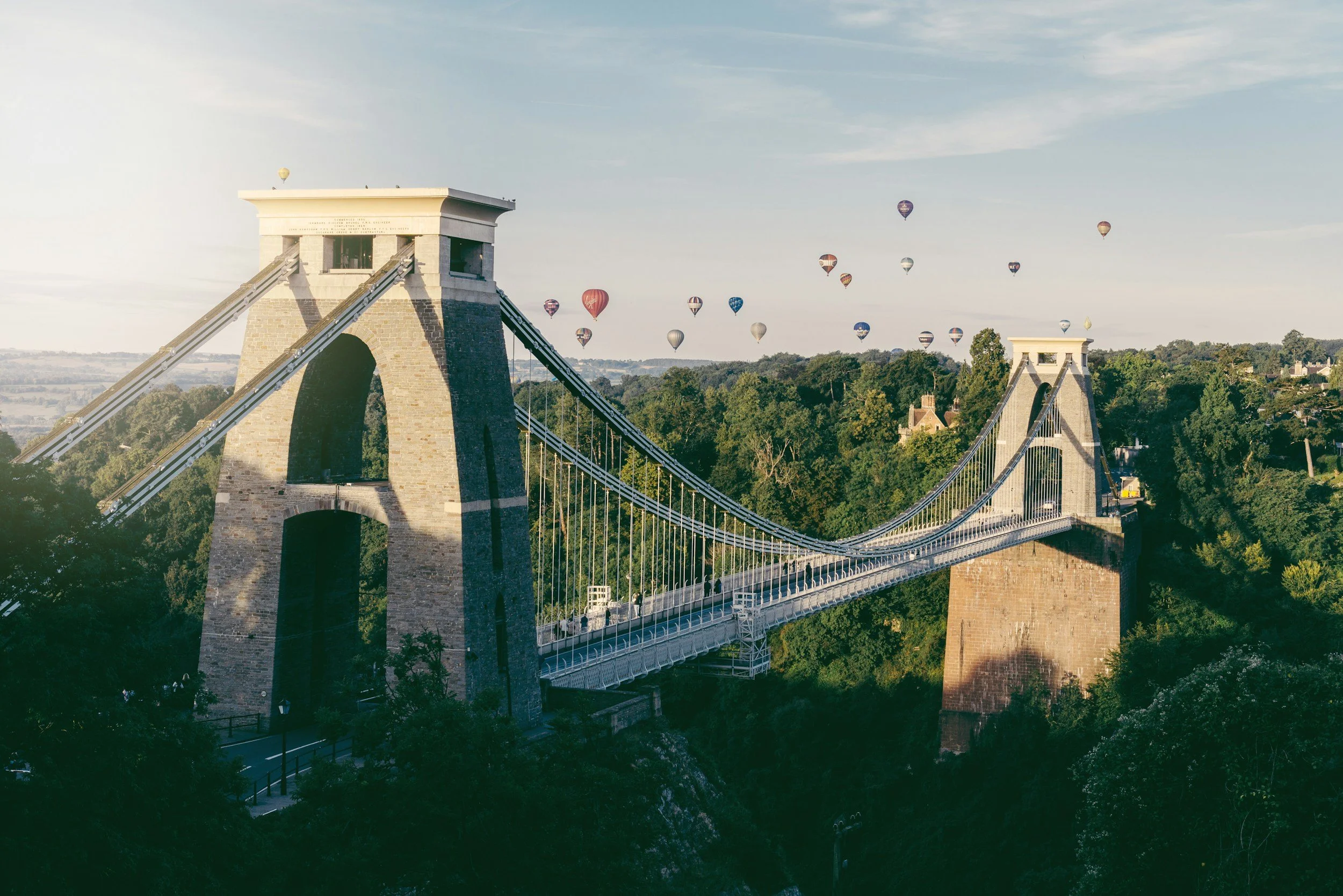 View of the Clifton Suspension Bridge with numerous hot air balloons floating in the sky above and greenery surrounding the bridge.