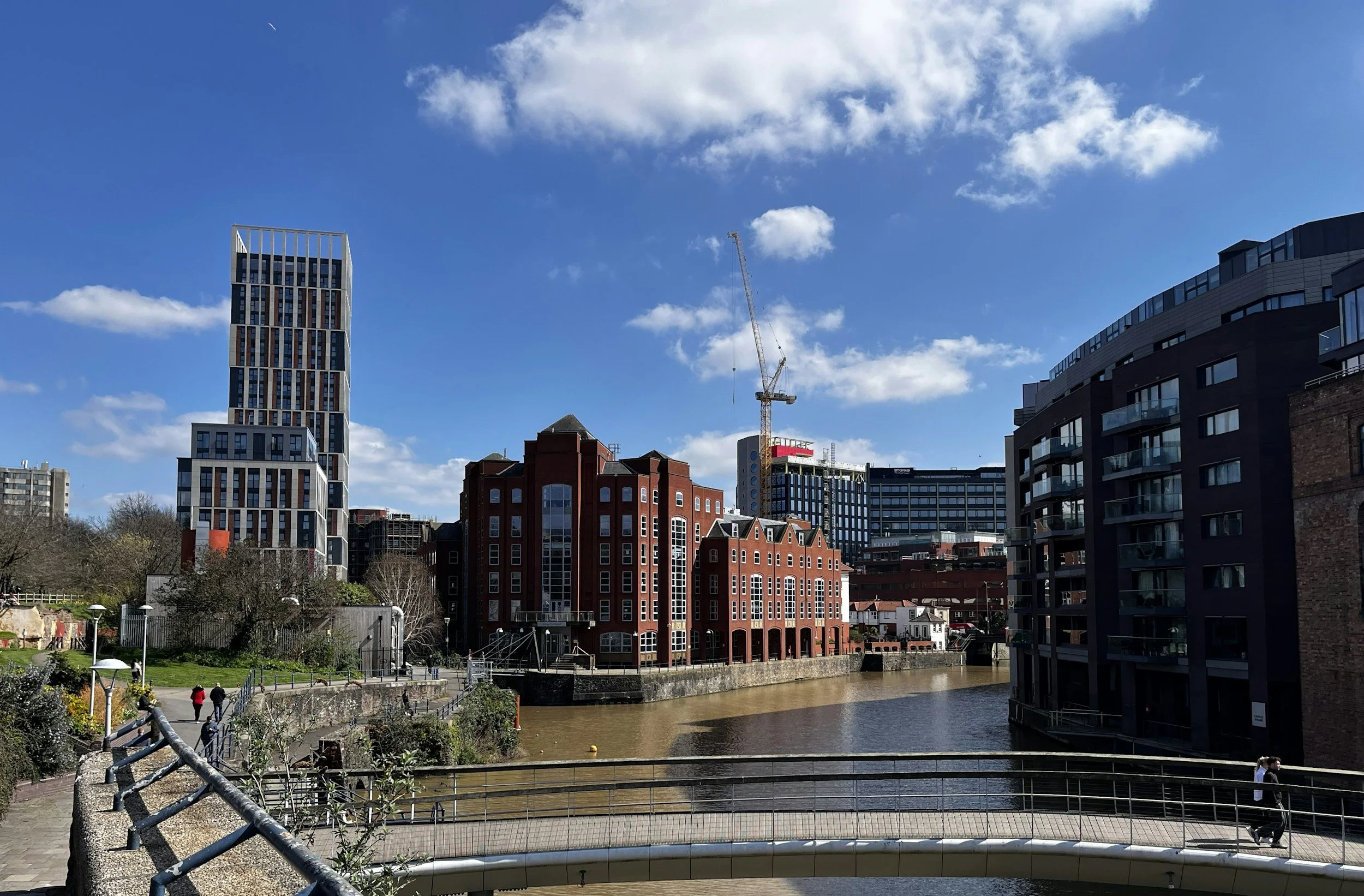 A cityscape with modern high-rise buildings and older brick buildings along a river, under a blue sky with scattered clouds.