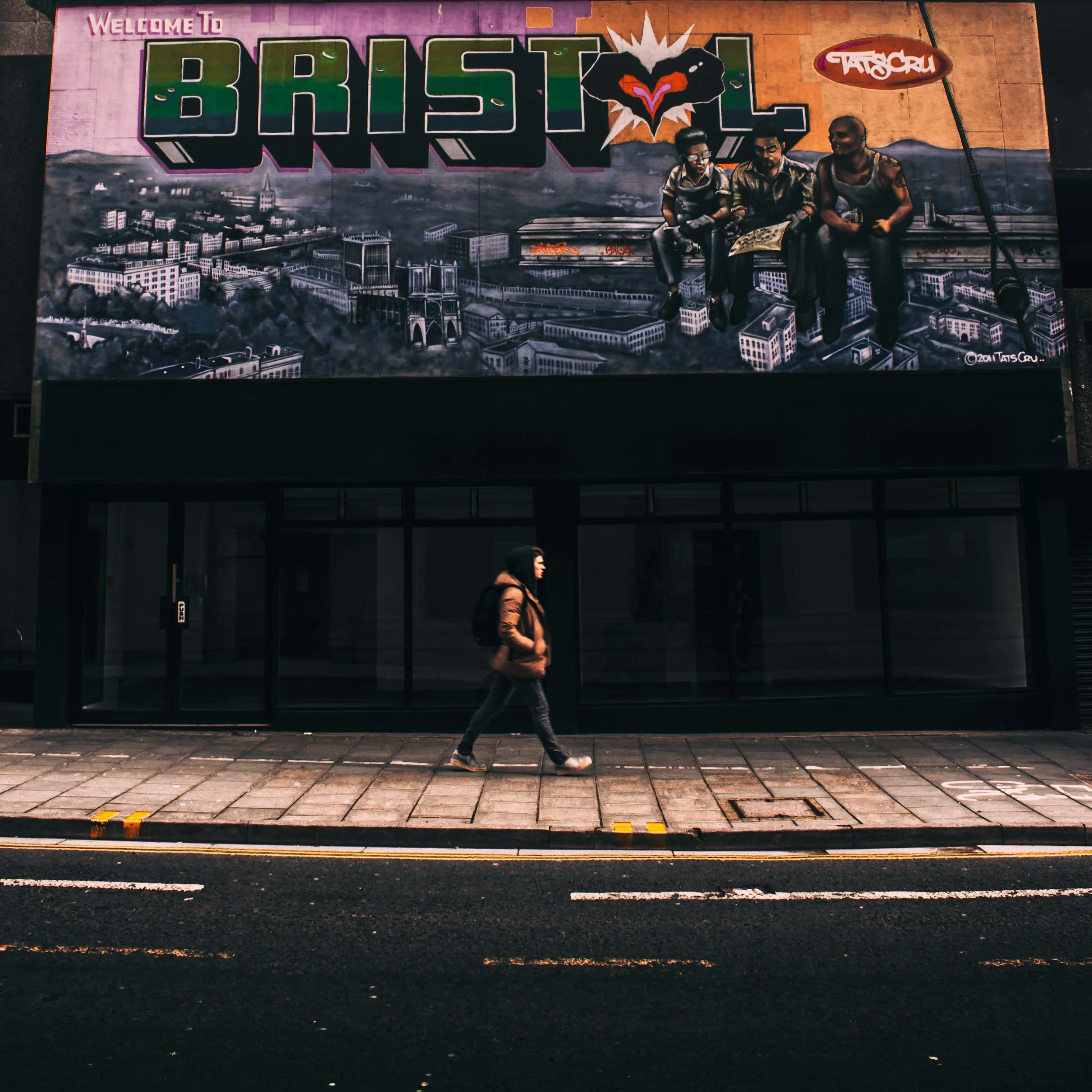Graffiti mural on a city wall that says 'Welcome to Bristol' with a black heart that has a pink and orange heart inside, and three people sitting on a ledge, with a cityscape background. A person walks in front of the mural. The scene is urban and artistic.
