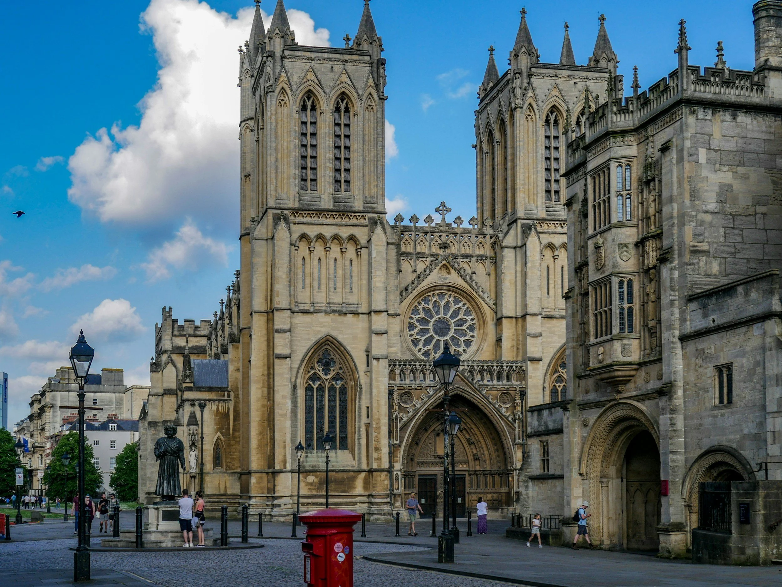 Front view of a historic Gothic-style cathedral with large stained glass windows, tall spires, and detailed stone architecture, with a partly cloudy blue sky in the background and people walking in the foreground.