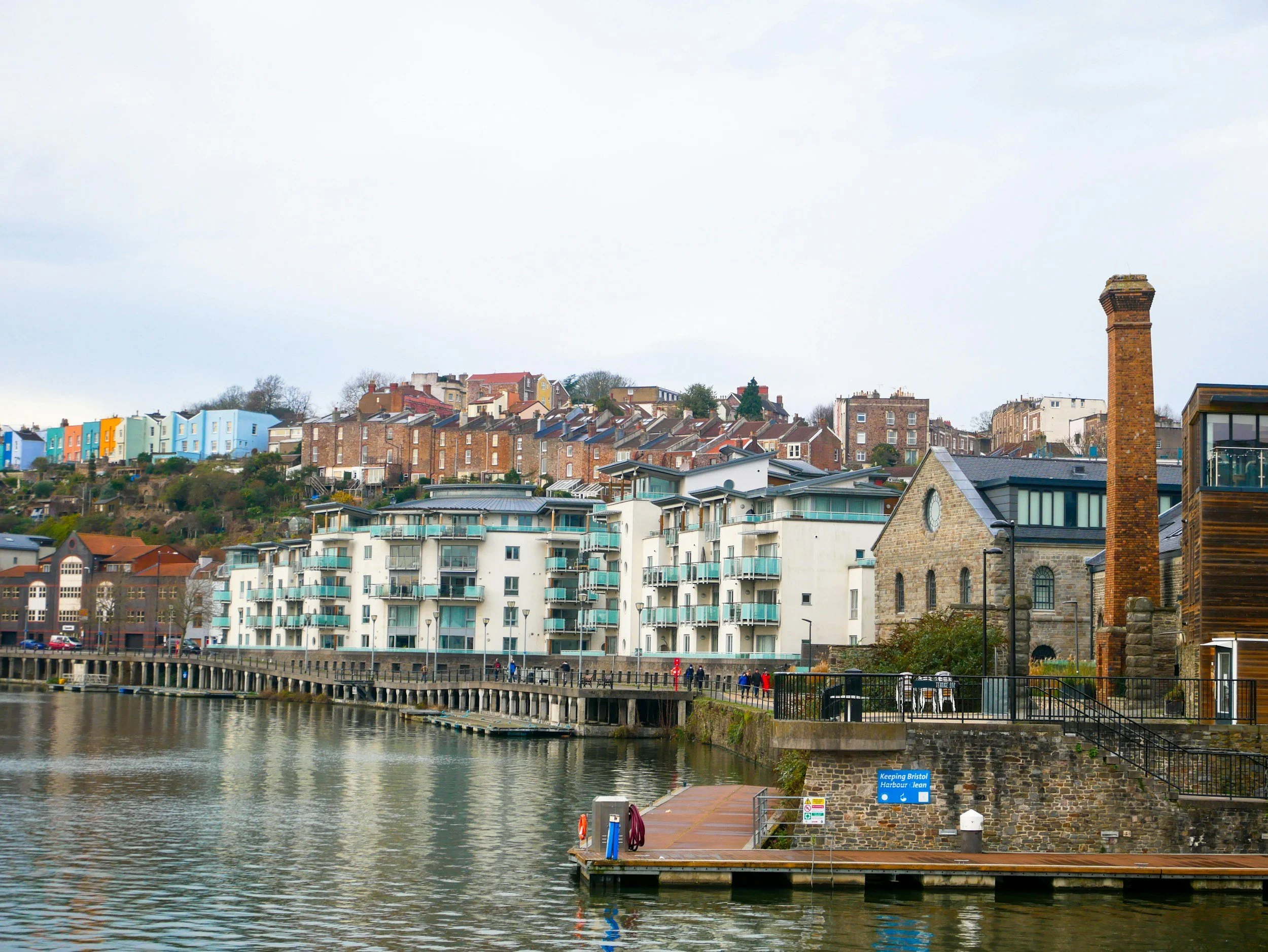 A waterfront scene with a buildings and houses perched on a hill, a stone building with a chimney in the foreground, and a wooden dock with a sign that reads "Keeping Bristol Harbour Clean" along the water.