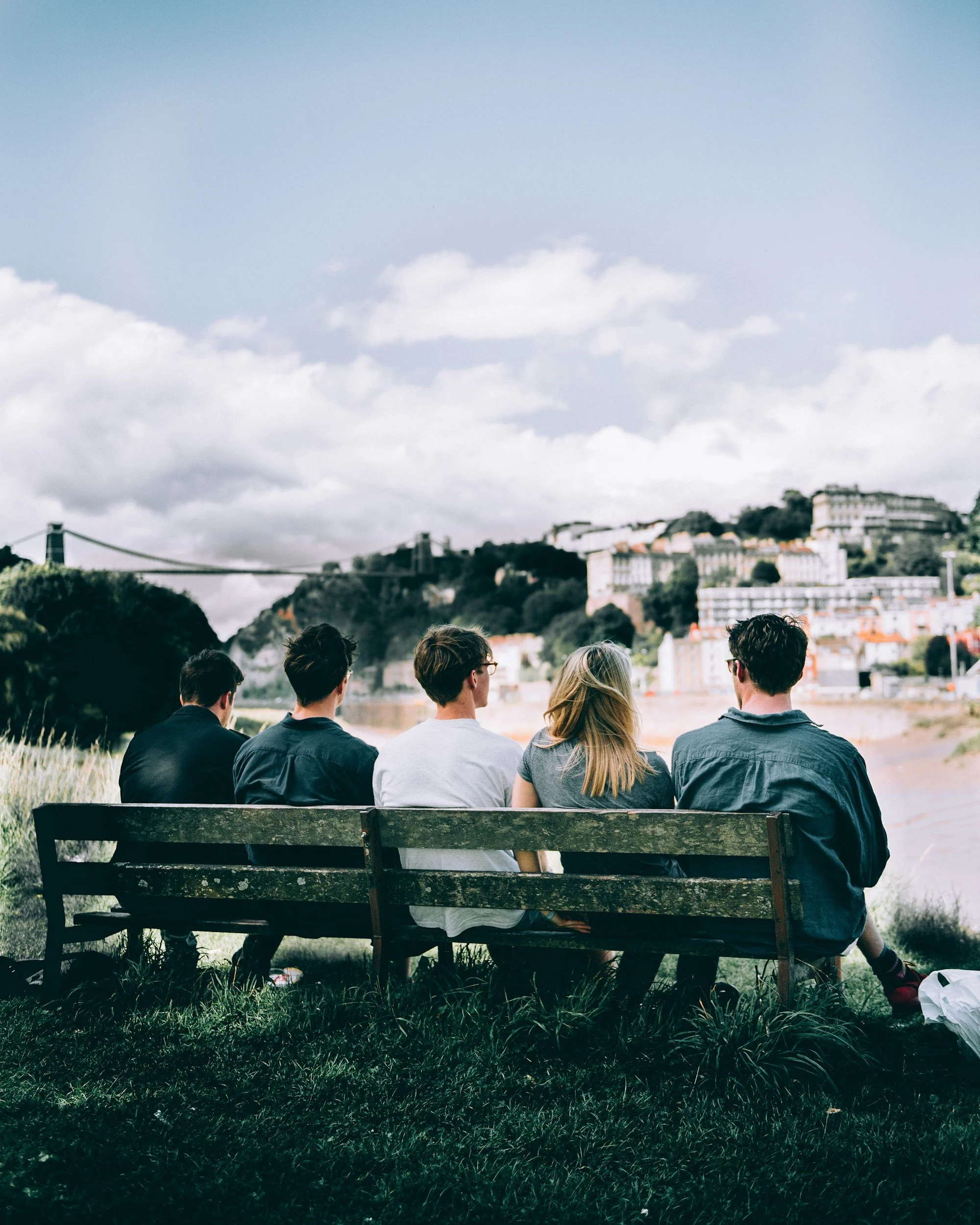 Group of young people sitting on a park bench facing a riverside with a city and hillside in the background, under cloudy sky.