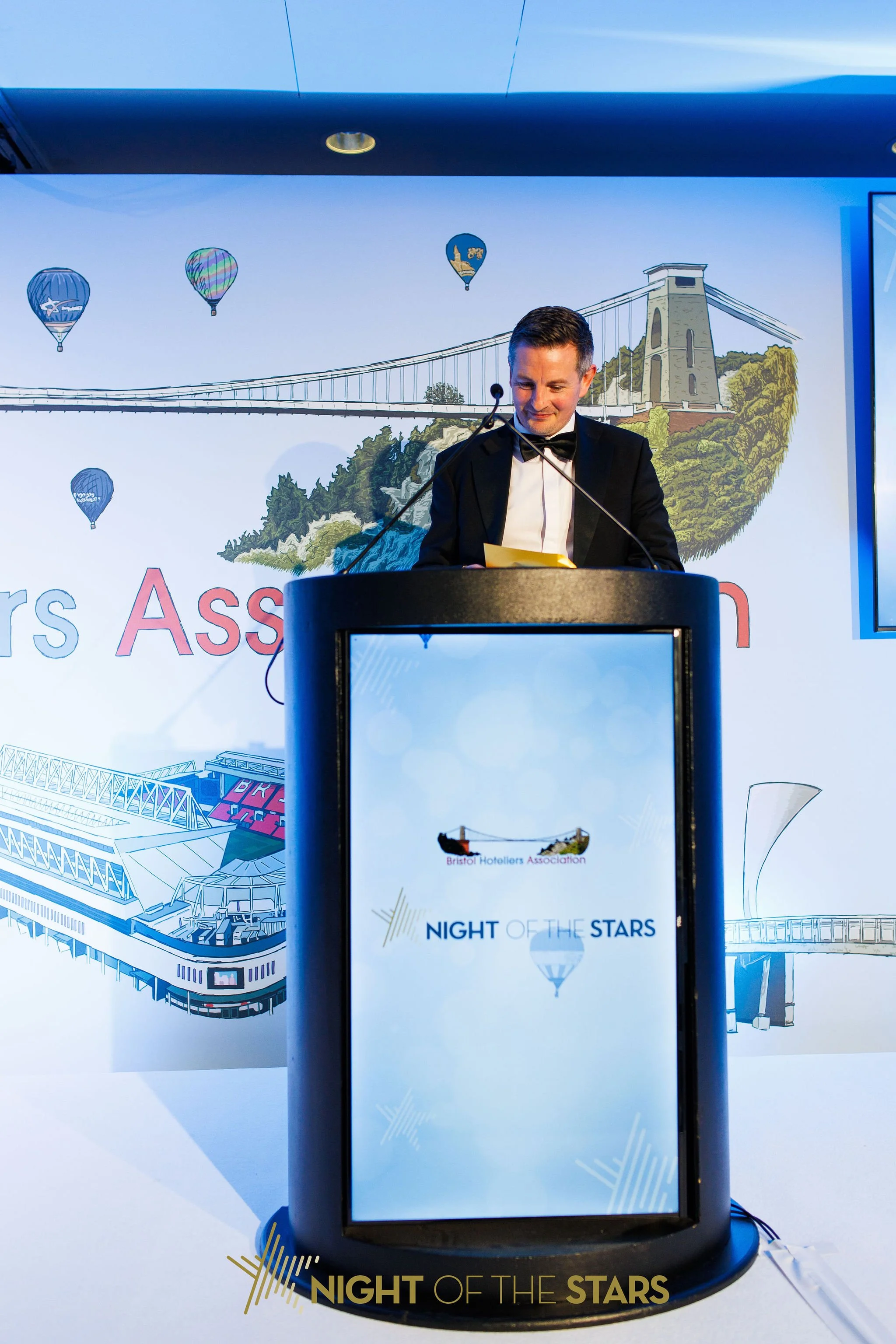 A man in a tuxedo stands behind a black podium with a glass front, reading notes at an event called "Night of the Stars" held by the Bristol Hoteliers Association. The backdrop features a scenic illustration of a suspension bridge, hot air balloons, 