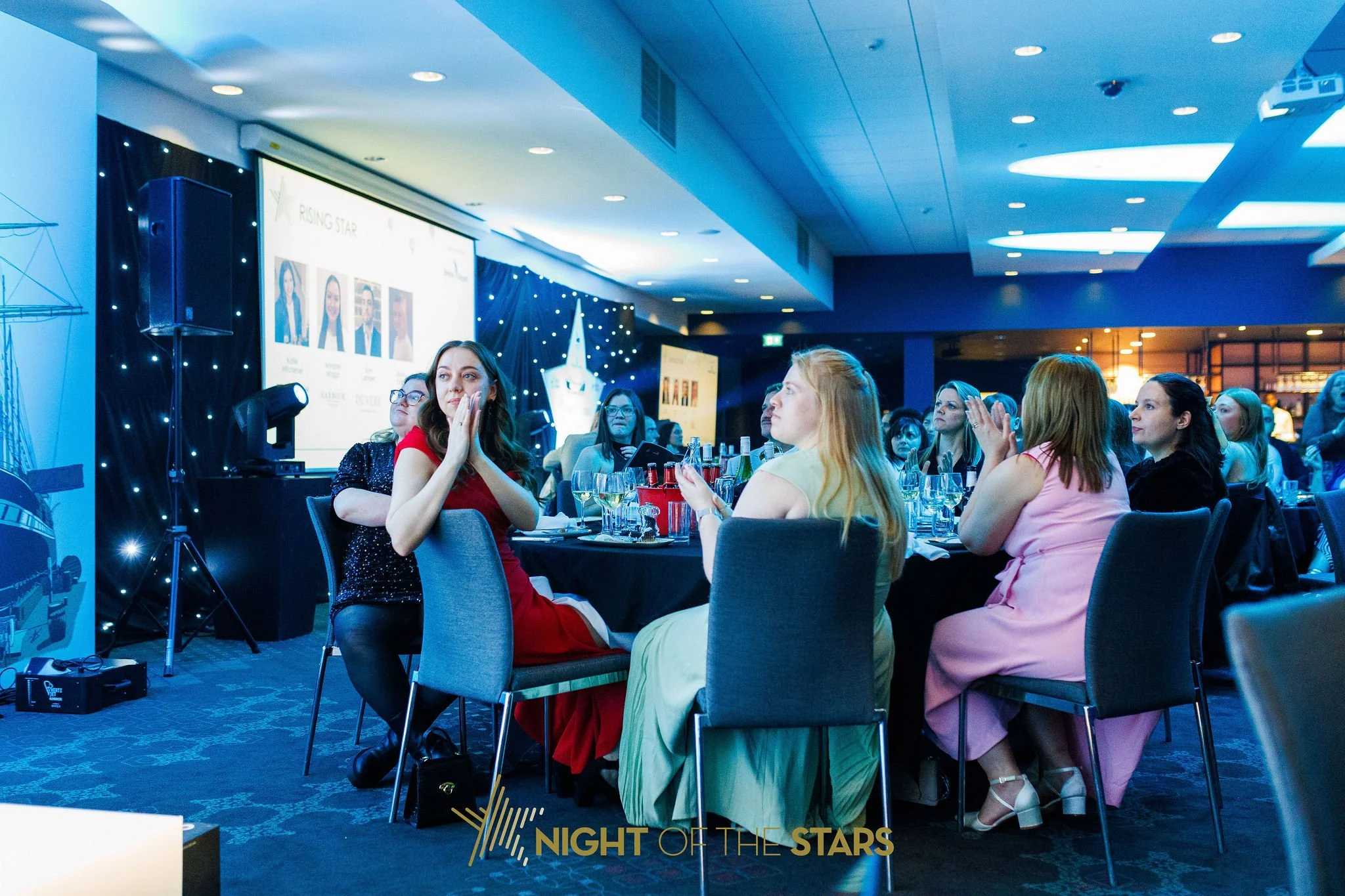 Indoor event with women seated at tables, some clapping, with a large screen in the background displaying photos of four women and event information, decorated with star and night sky themes.