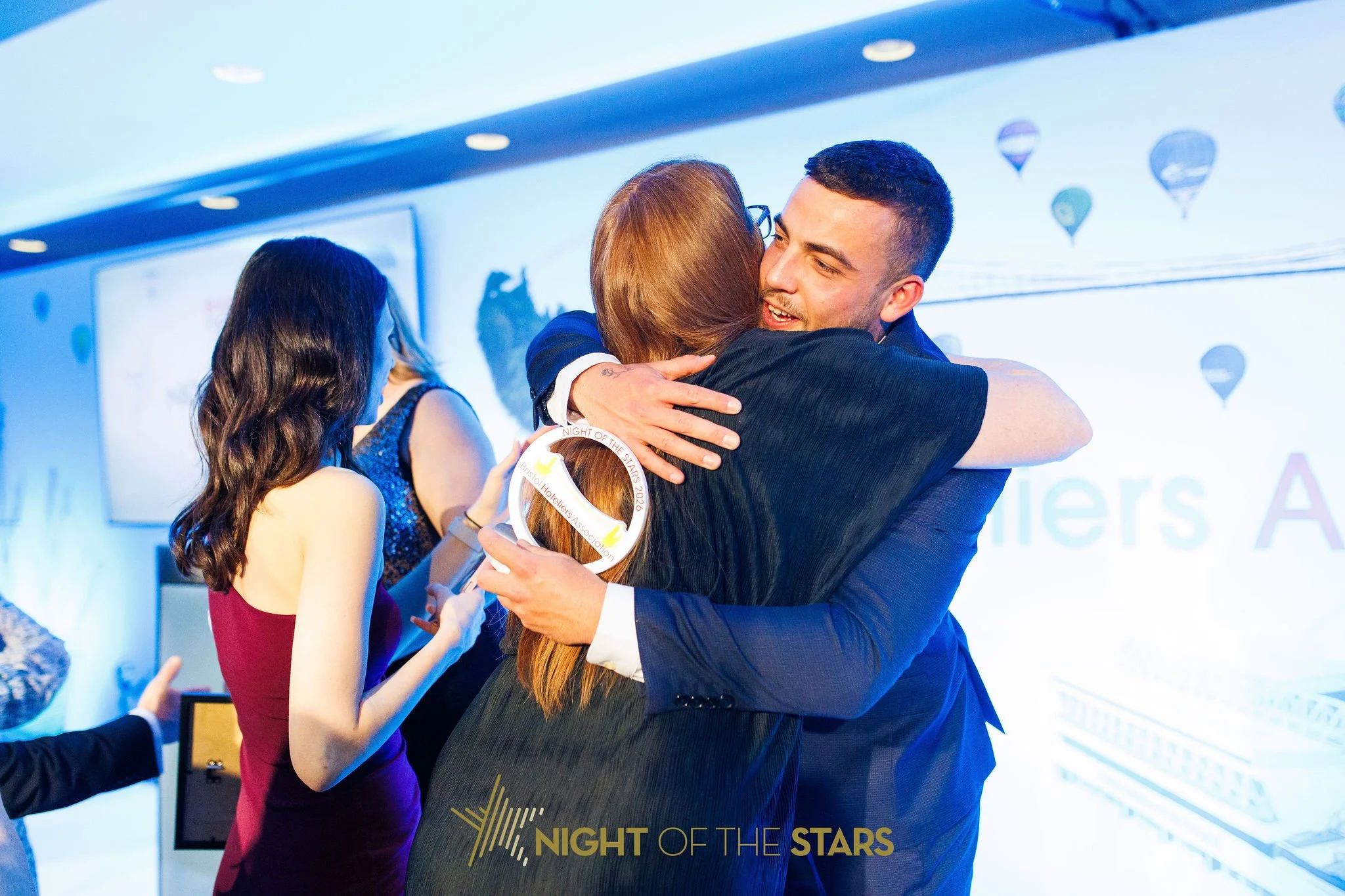 A man and a woman embracing each other at an awards event, with the woman holding a circular award. Other people are in the background, and the event appears to have a 'Night of the Stars' theme.