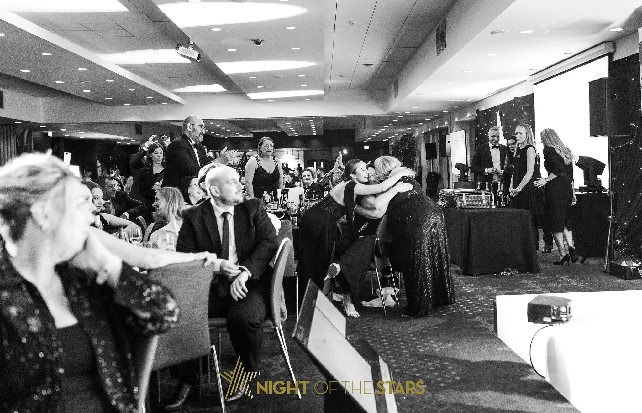 People attending a formal event or awards ceremony, with some hugging, speaking, and mingling, in a well-lit banquet hall with a stage on the right.