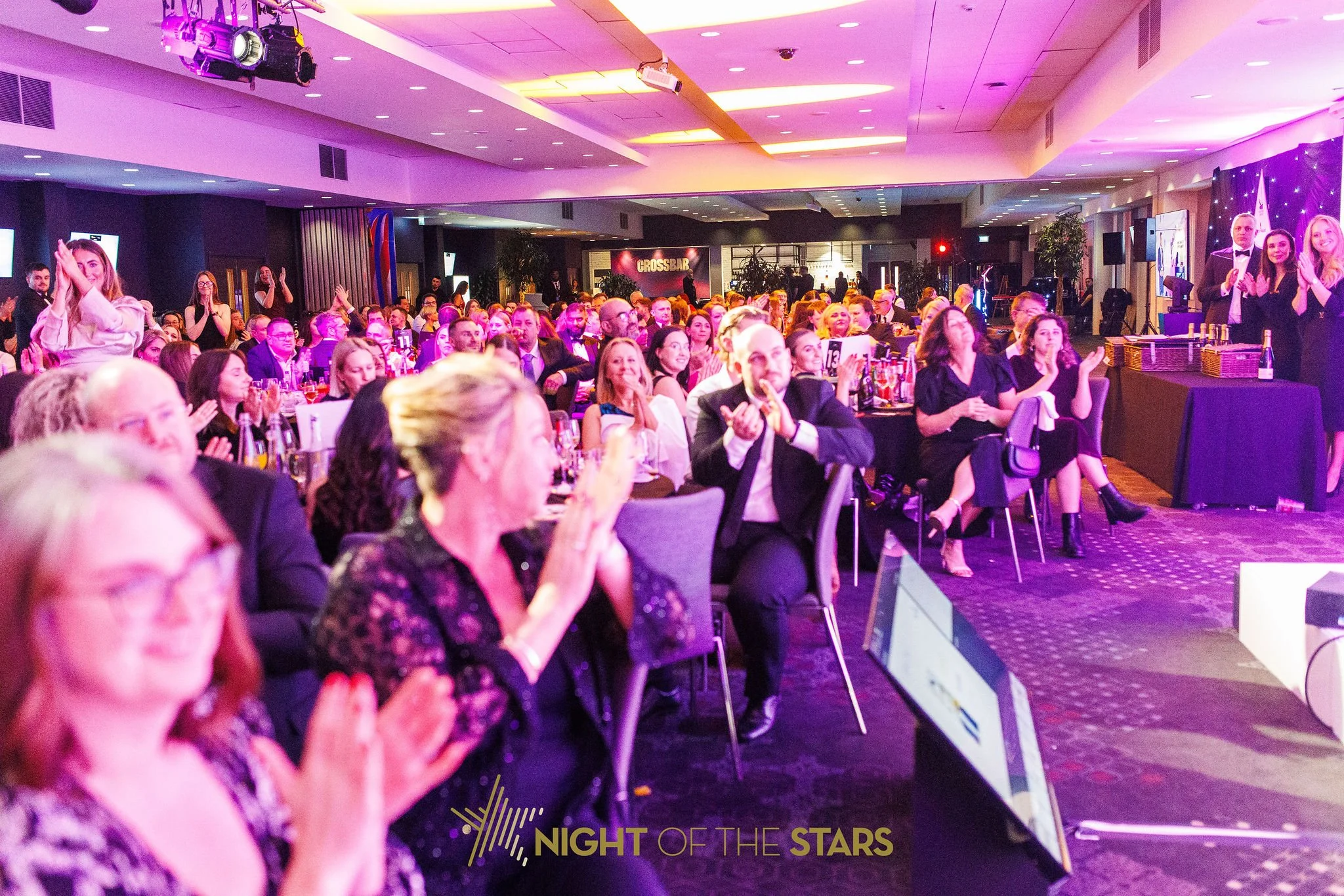 Crowd of people dressed in formal attire at an indoor event with purple and pink lighting, a stage area on the right side, and a banner that reads 'Night of the Stars' in the lower part of the image.