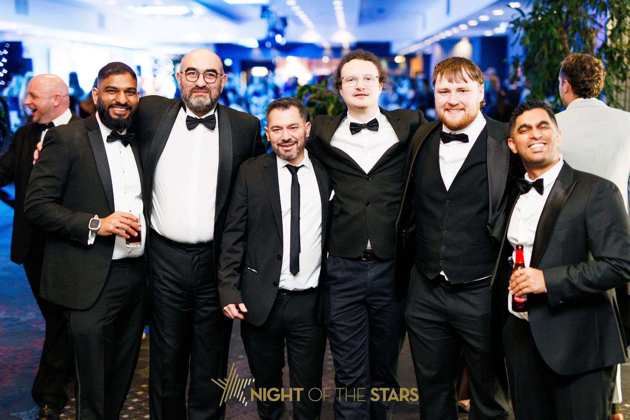 Group of six men dressed in tuxedos at a formal event, smiling and posing for the photo in a venue decorated for 'Night of the Stars.'