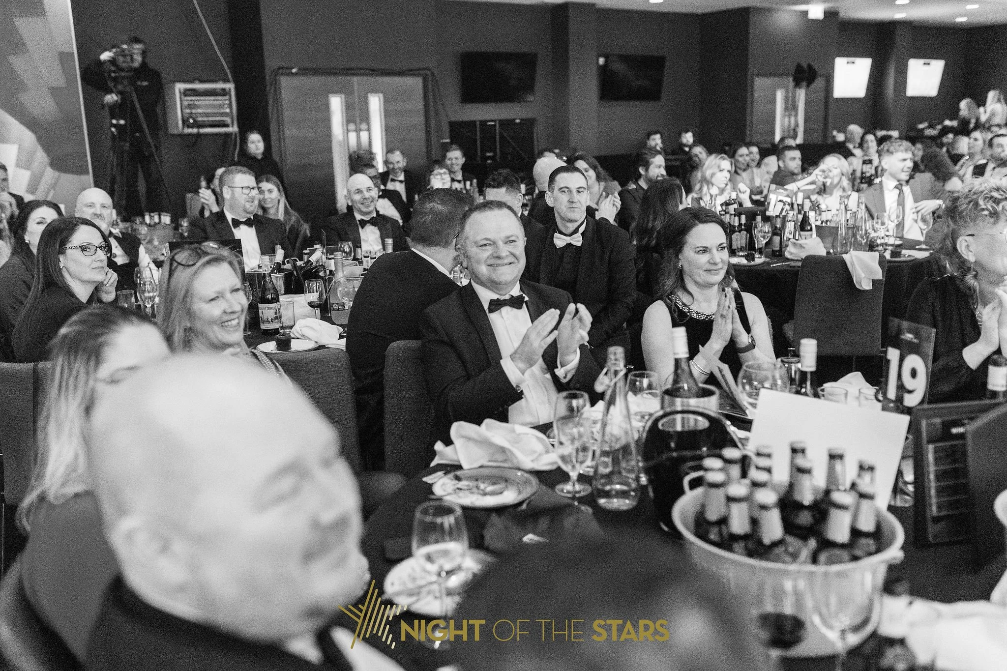 A group of people dressed in formal attire, sitting at round tables, attending a formal event or banquet. Some individuals are clapping and smiling, with wine glasses and bottles on the tables. The background shows a dark room with multiple TV screen