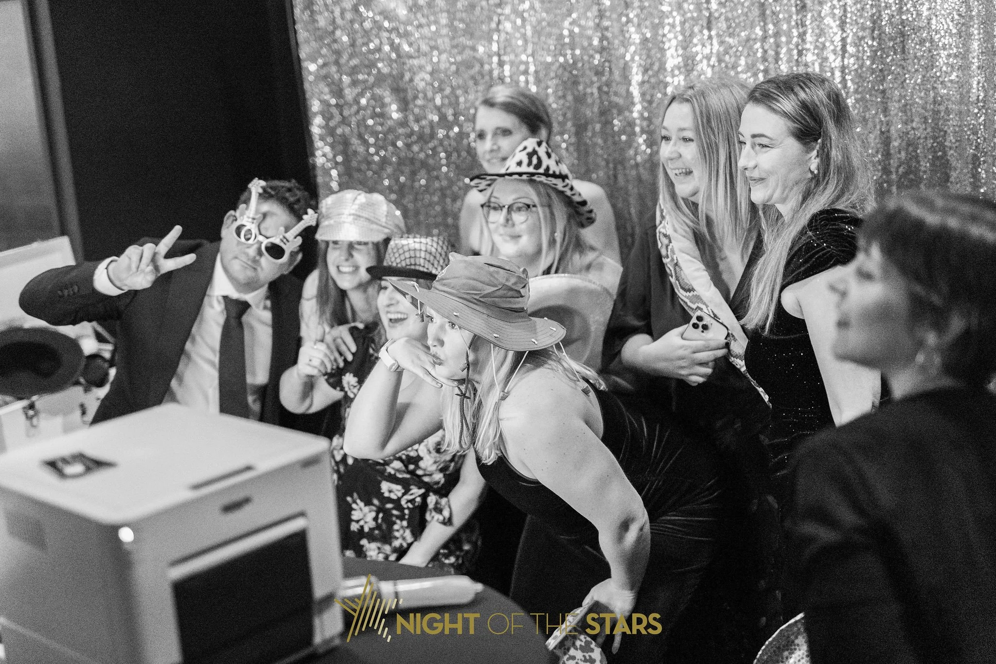 Group of people at a celebration taking a selfie, dressed in party costumes and accessories, with a sparkly backdrop and a "Night of the Stars" sign.
