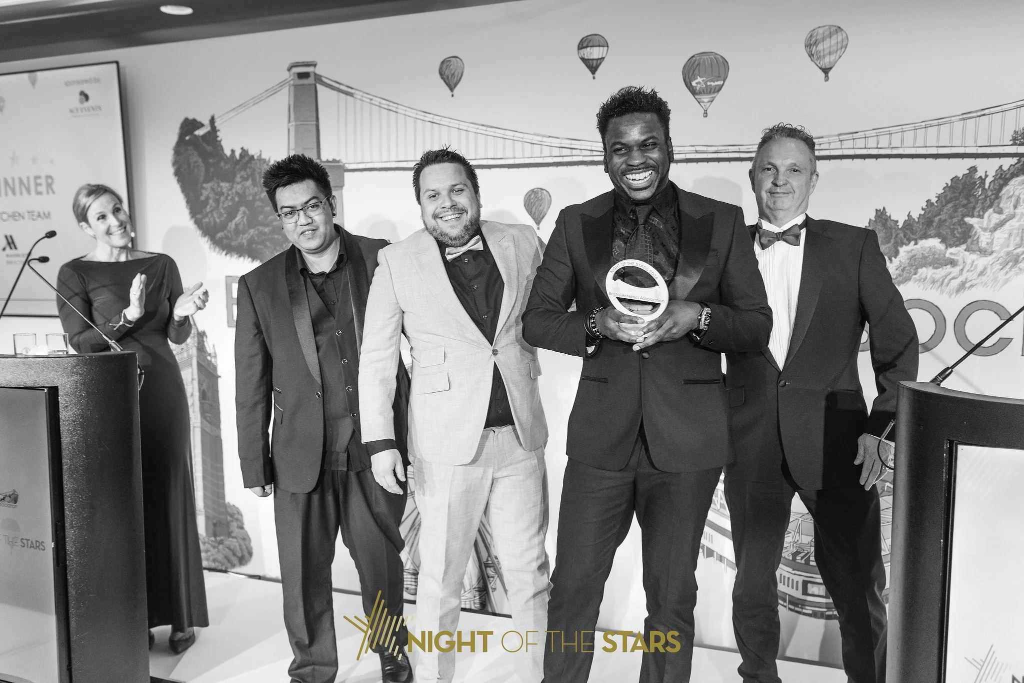 Group of five professionally dressed people standing on stage at an awards event, with a backdrop showing hot air balloons and a bridge, holding a trophy and smiling.