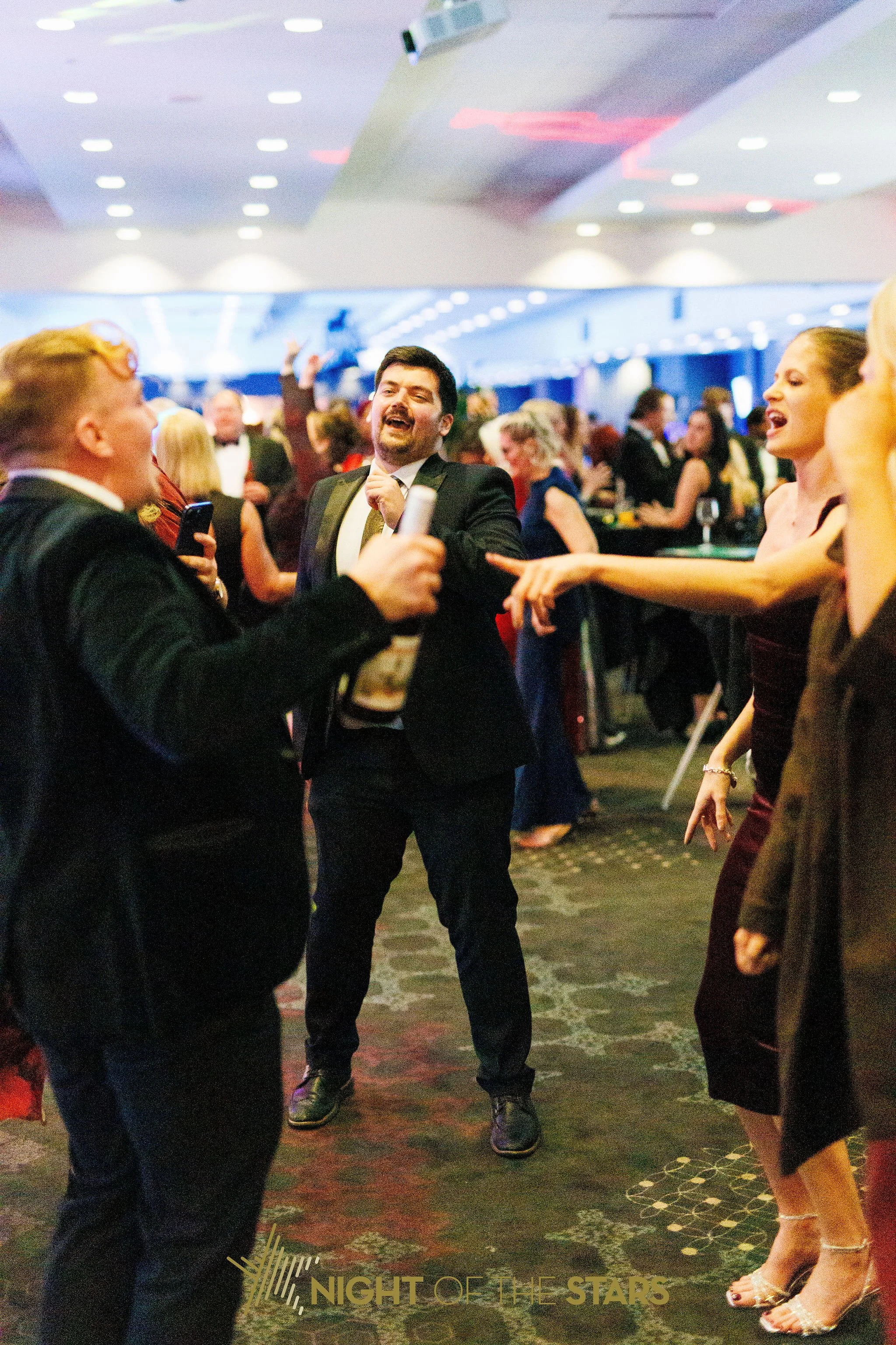 People dancing and socializing at a formal event called 'Night of the Stars' in an elegant indoor venue with patterned carpet and dim lighting.