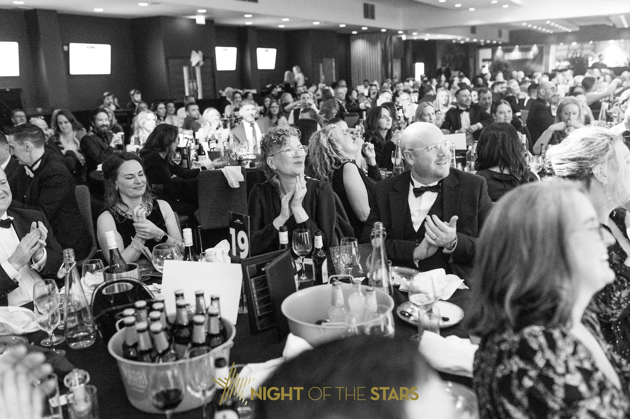 Black and white photo of a large formal gathering at an event in a banquet hall, with guests seated at tables dressed in black-tie attire. People are smiling and engaging, with many holding glasses or food. The tables have bottles, glasses, and decorations. The background shows more guests and bright screens or displays.