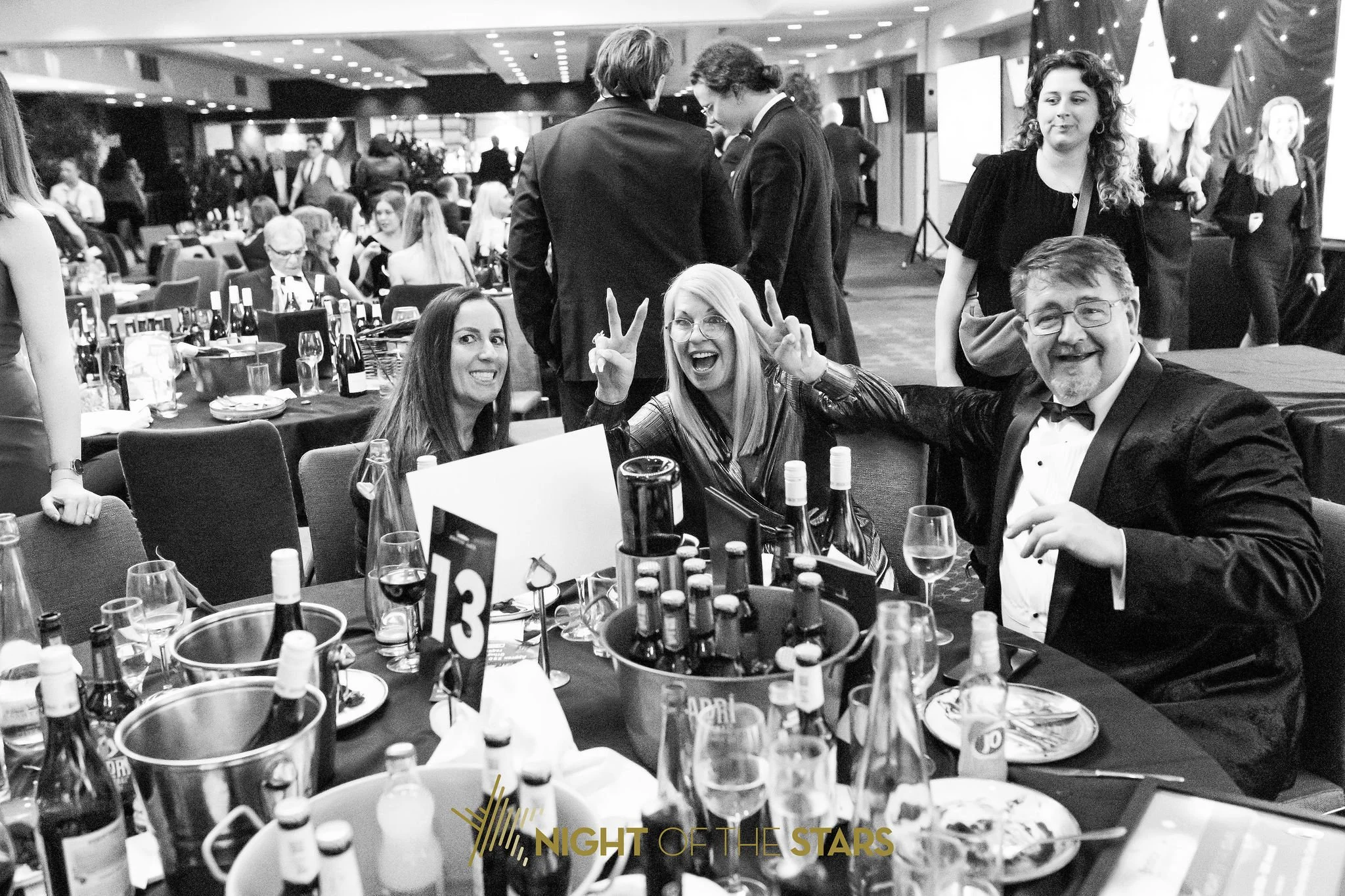 People celebrating at a formal event or dinner in a large banquet hall, with tables set with bottles of wine and glasses, and some individuals posing and smiling at the camera.