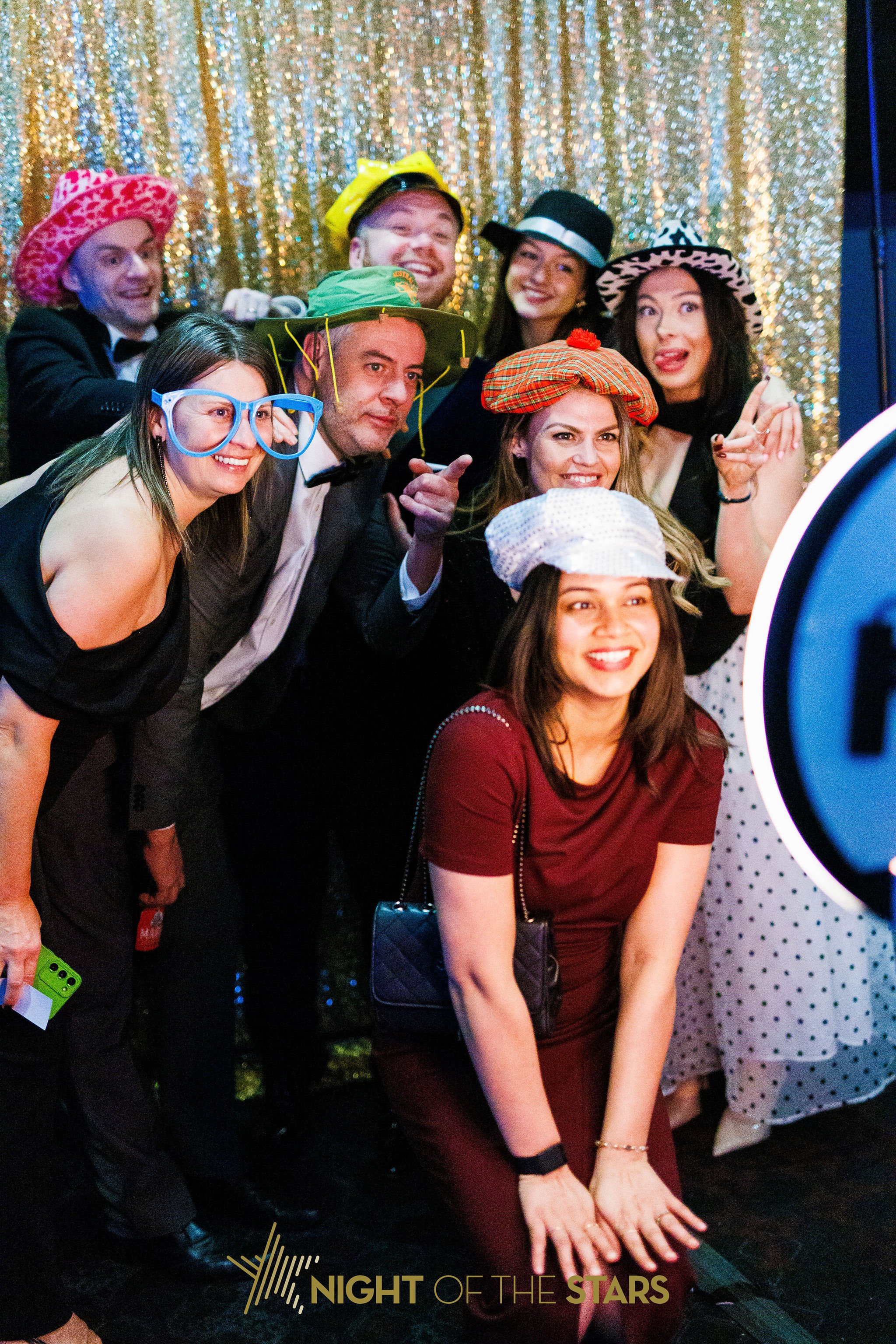 Group of people dressed in colorful and whimsical costumes and hats, posing for a photo in front of a shiny, gold backdrop at an event titled 'Night of the Stars'.
