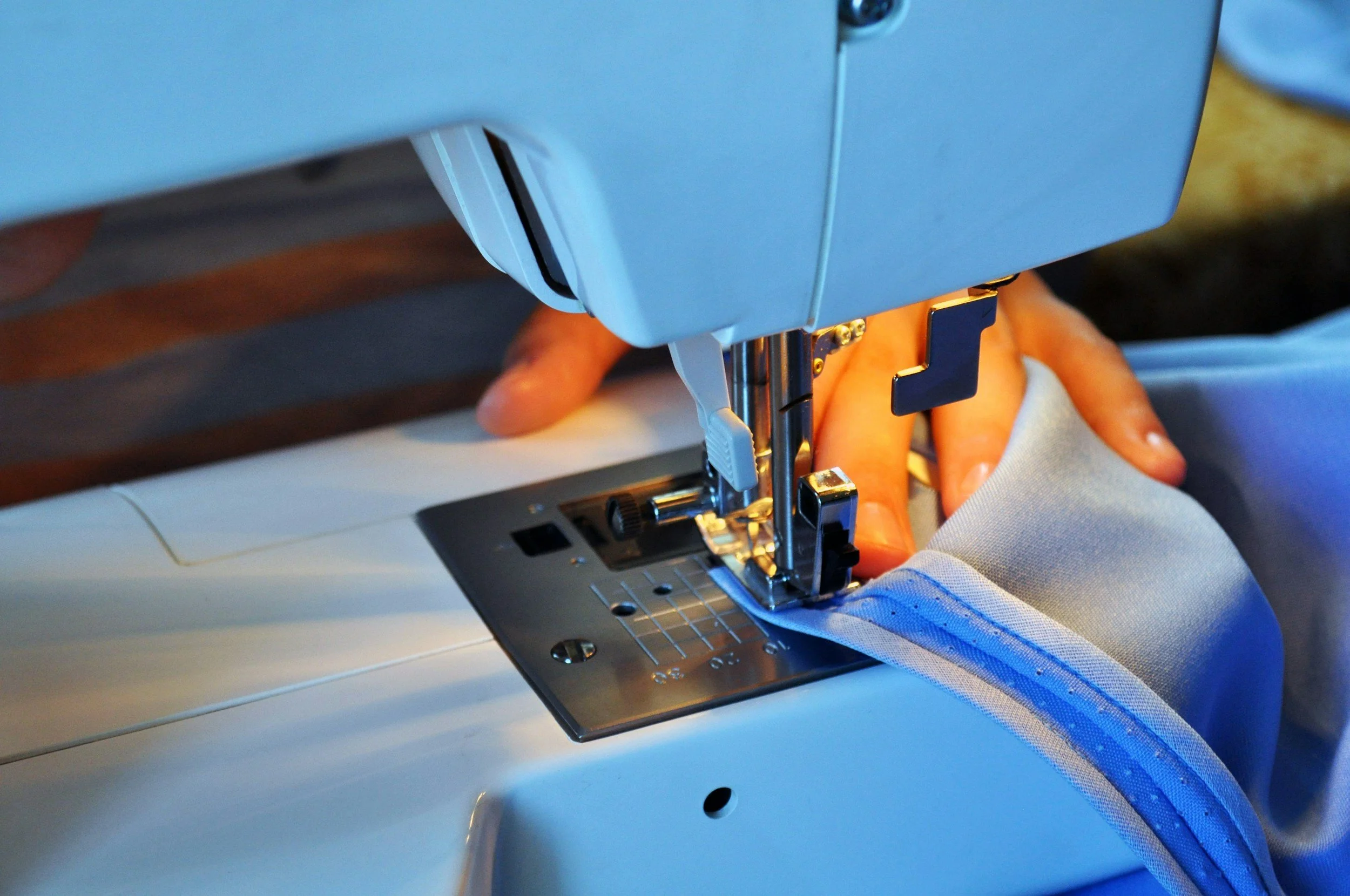 Close-up of a person's hands guiding light blue MB Sport fabric through a white sewing machine.