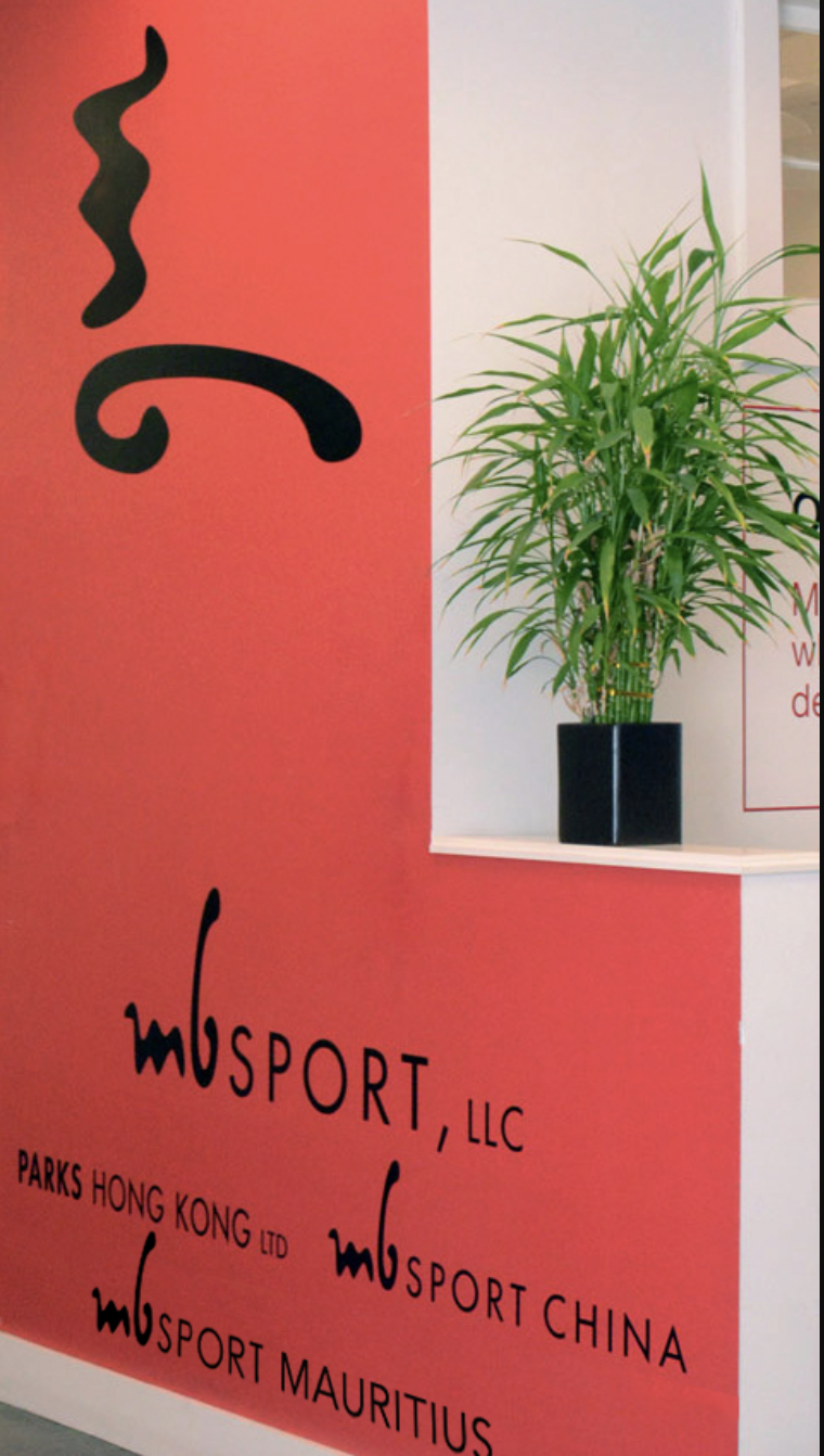 Red wall with black text and MB Sport logo, next to a potted green plant placed on a white surface.