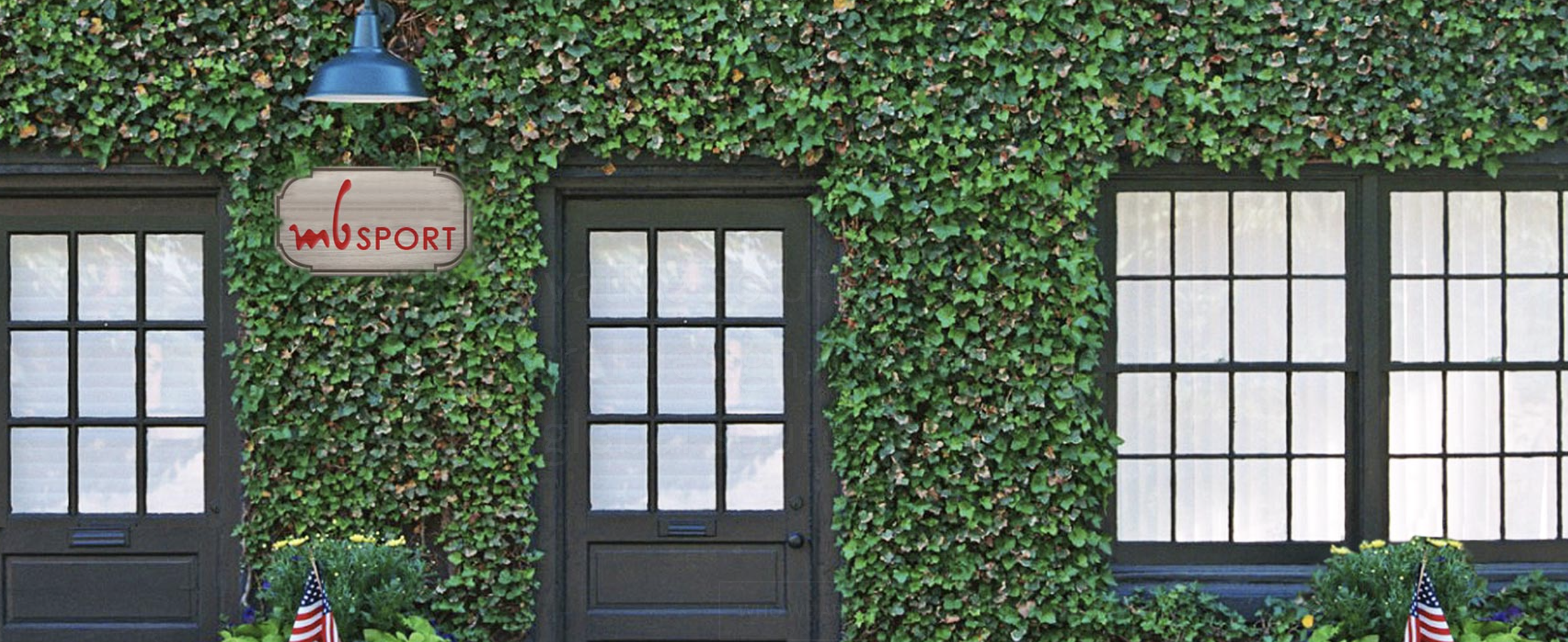 Building with black-framed windows and doors, covered in green ivy, with a sign reading "MB SPORT" and American flags in front.