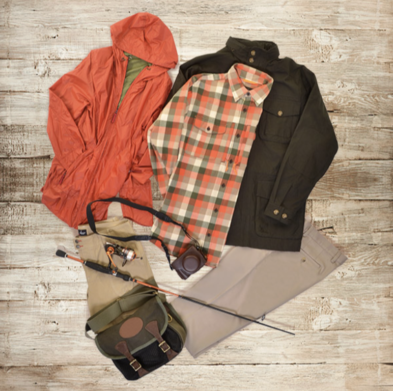 Outdoor clothing and gear laid out on a wooden surface, including a red rain jacket, a plaid shirt, a black jacket, khaki pants, a fishing rod, and a small backpack.
