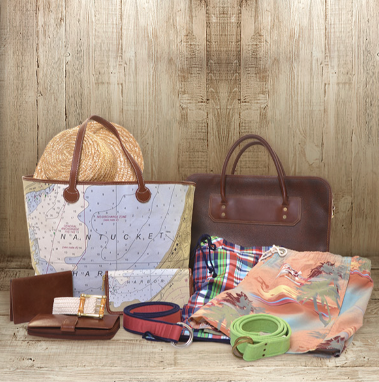 Travel essentials including a straw hat, a map-themed tote bag, a brown leather bag, a colorful plaid shirt, a floral scarf, two belts, and a wallet and belt on a wooden surface against a wooden background.