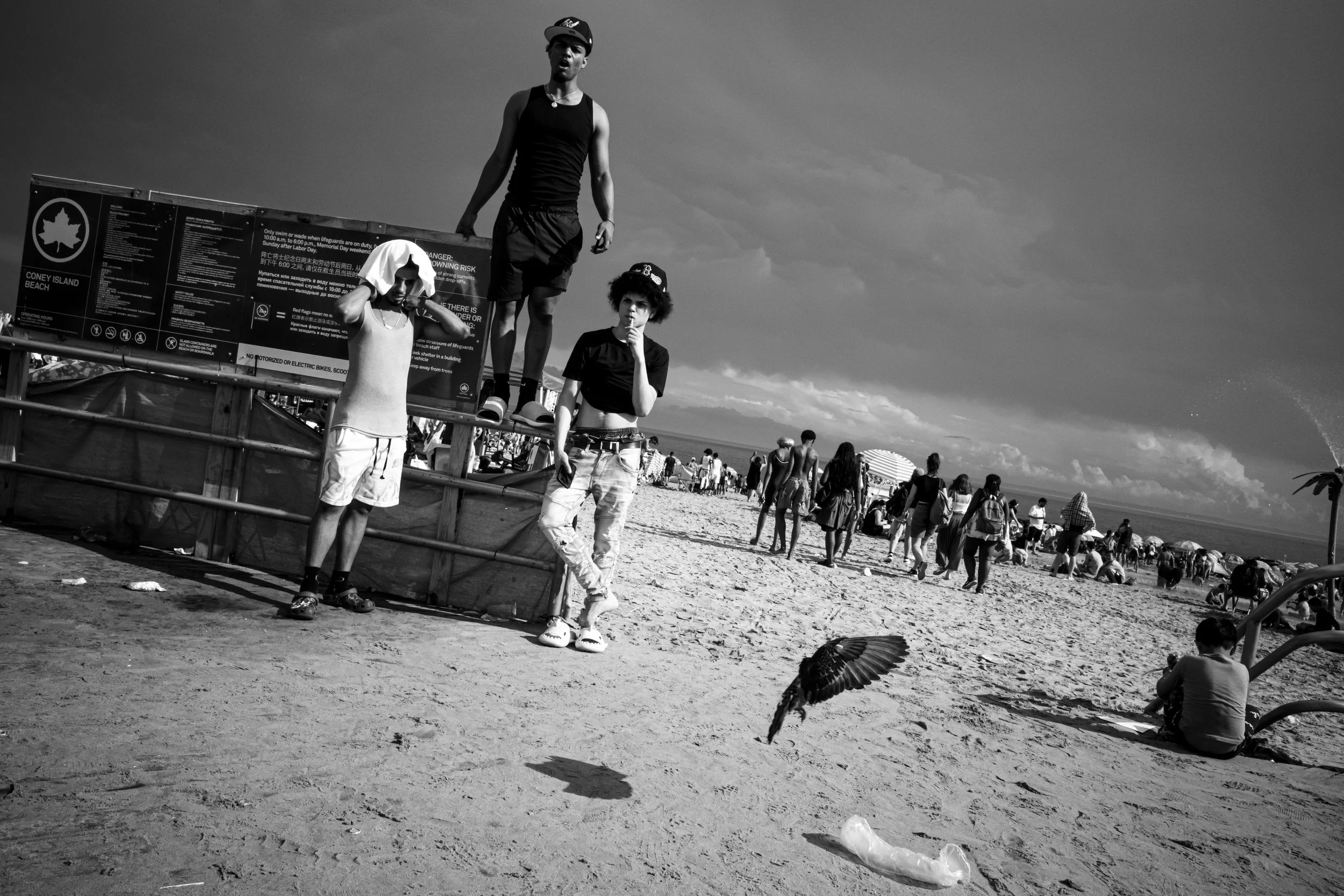 Black and white photo of people at a crowded beach. Three people stand on a wooden structure, with one person holding a shoe to their face, another with arms crossed, and a third adjusting their hat. Many beachgoers are in the background, some sittin