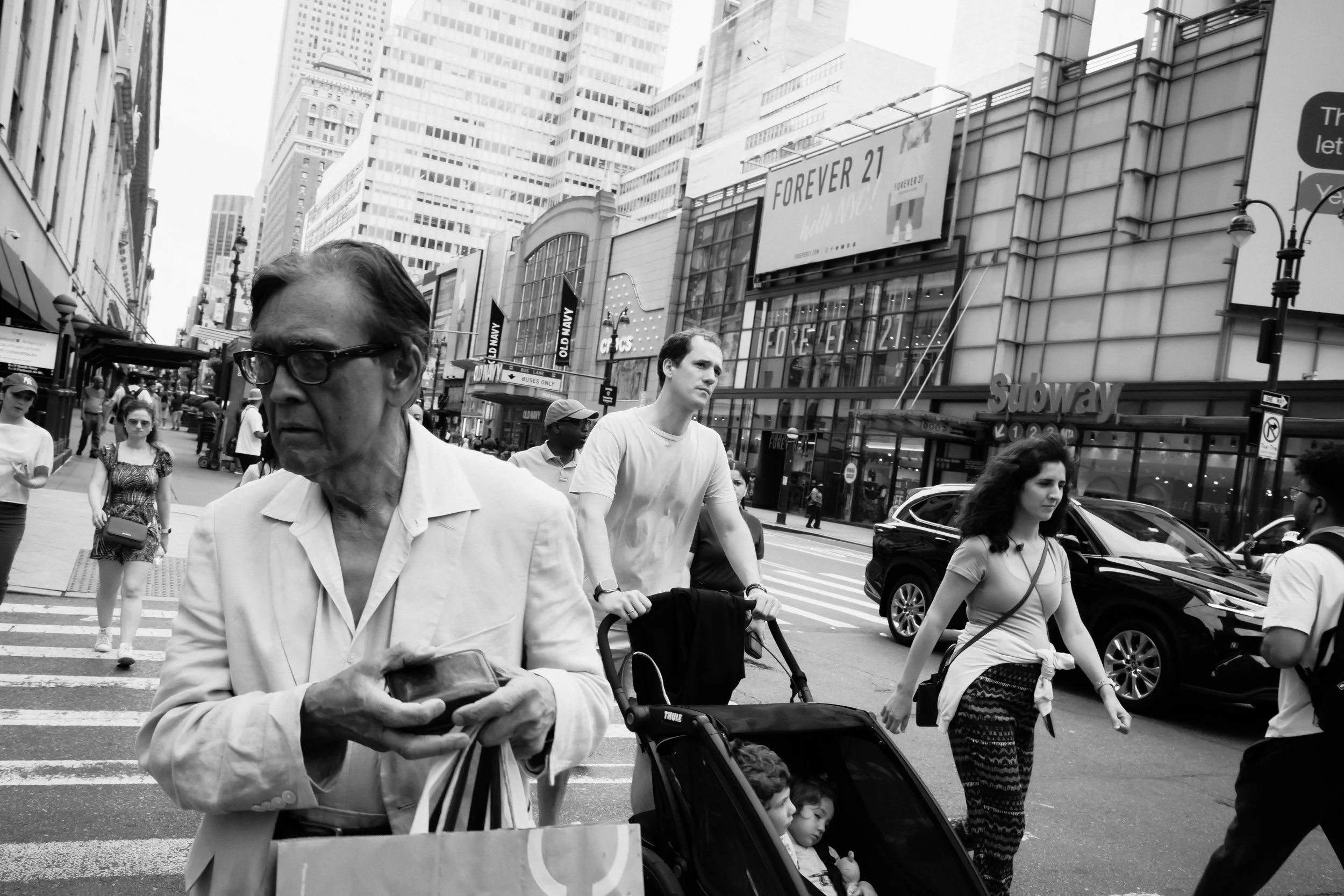 People crossing a city street in front of tall buildings with billboards. Foreground features an older man with glasses, and a woman pushing a stroller with two children. Other pedestrians walk along the sidewalk.