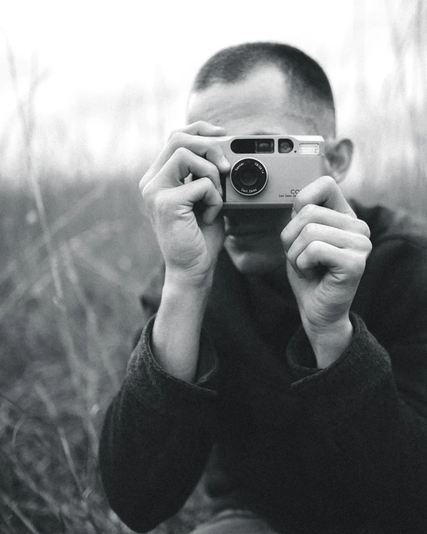 A person with short hair taking a selfie with a vintage camera in black and white, standing outdoors in tall grass.