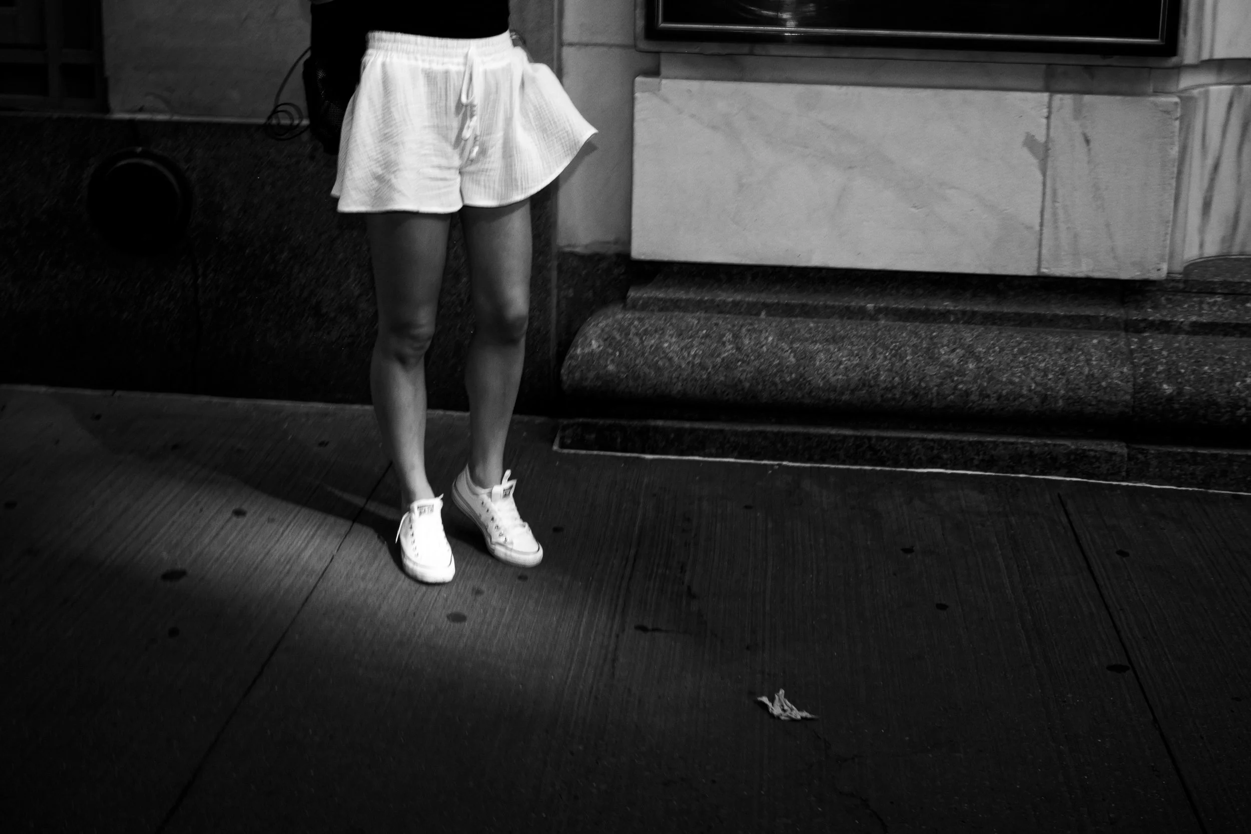 The image shows the lower half of a person standing on a sidewalk. They are wearing white shorts, white sneakers, and a black top. The background includes a stone wall, a window, and some objects on the sidewalk, including a small piece of trash.