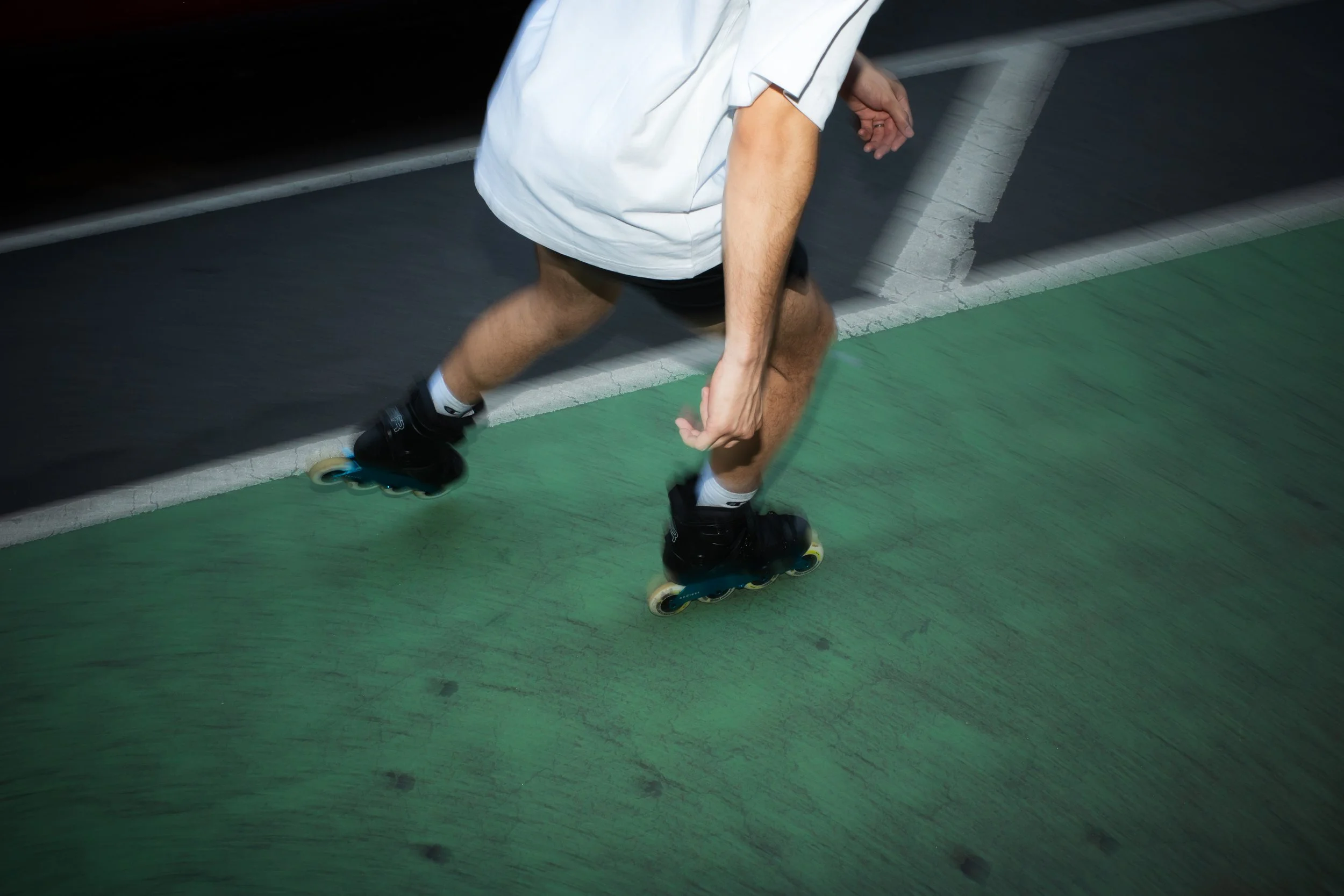 A person roller skating outdoors on a green and black surface, wearing black roller skates, black shorts, a white shirt, and white socks.