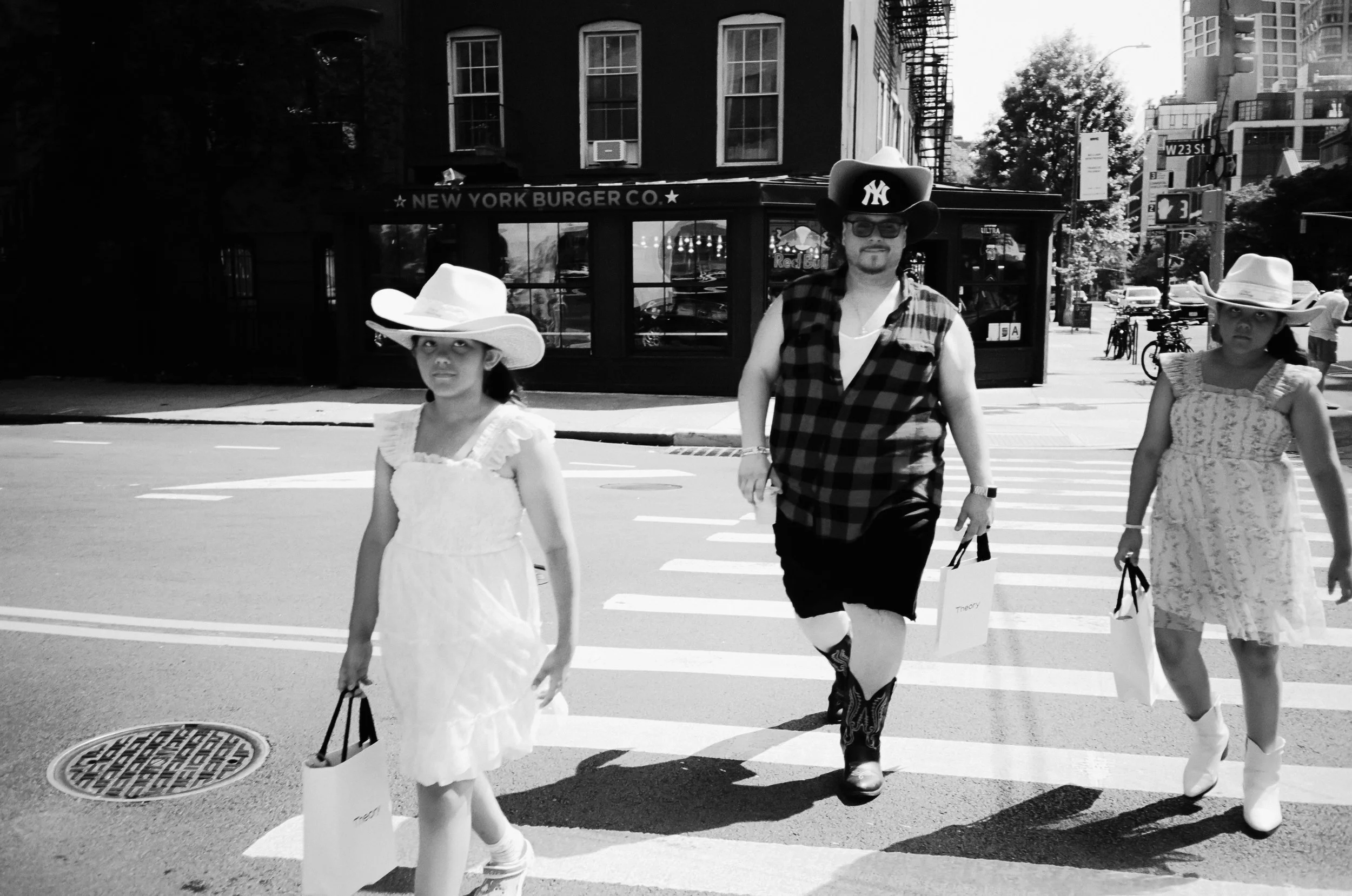 A black-and-white photo of a man walking across a crosswalk with two young girls. The man is wearing a sleeveless plaid shirt, cowboy boots, sunglasses, and a large cowboy hat with a New York Yankees logo. The girls are dressed in dresses and wearing