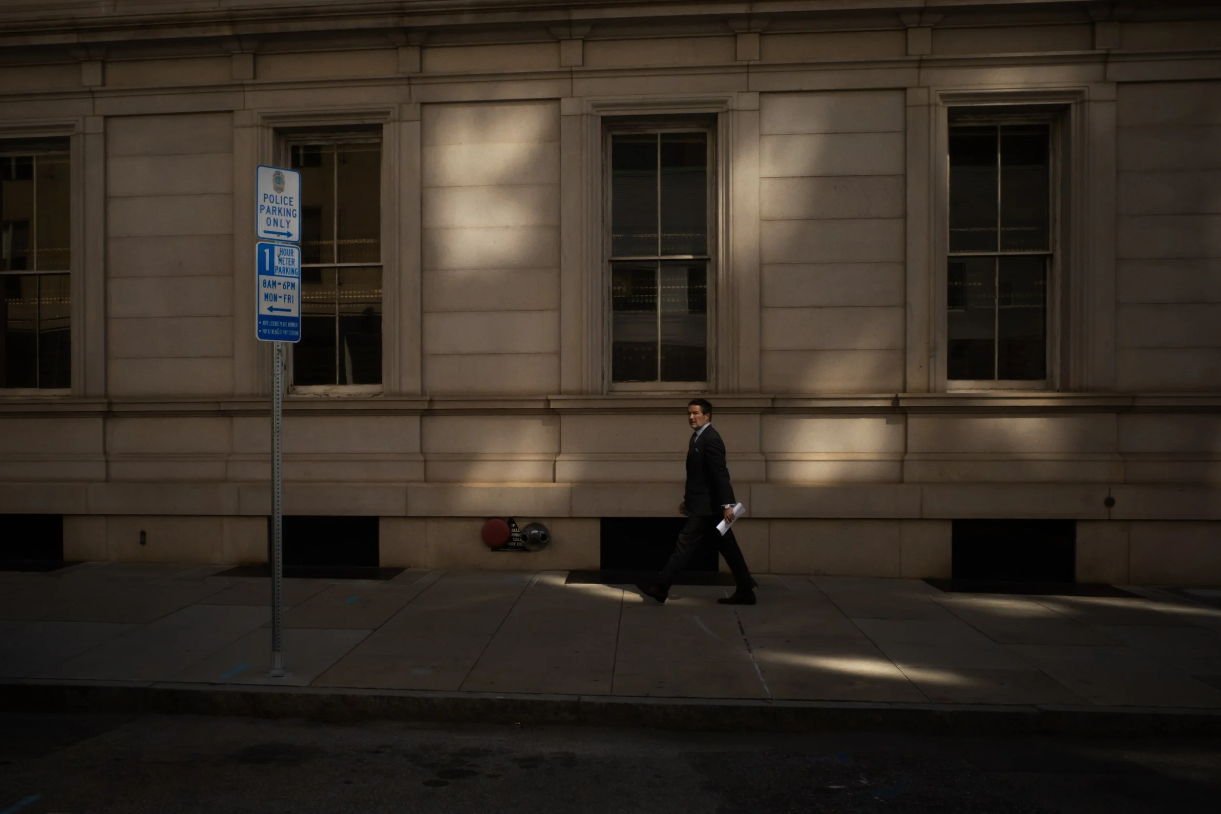 A man in a suit walking on the sidewalk in front of a stone building with three large windows. There is a police parking only sign on a pole near the sidewalk.