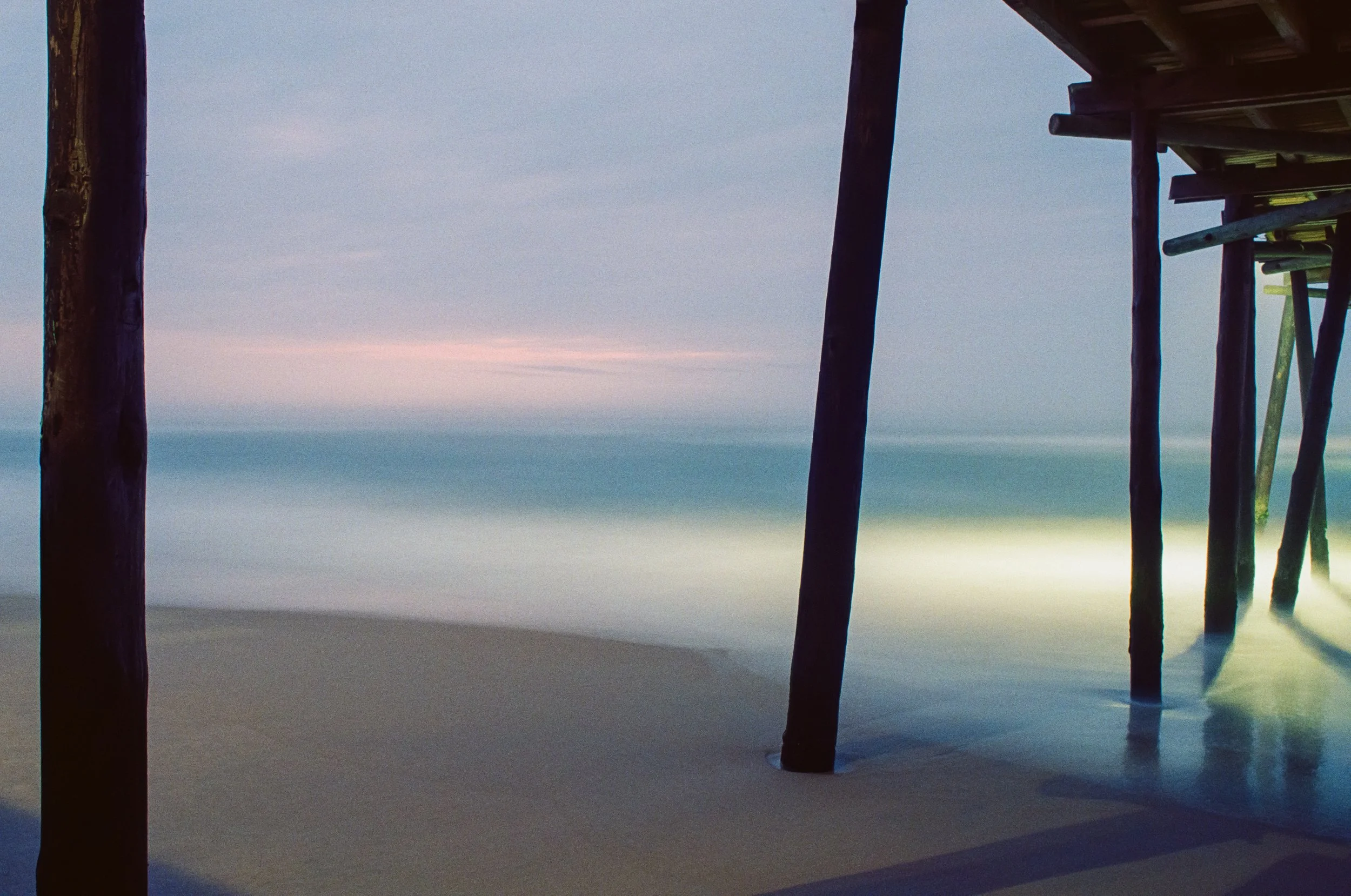 Underwater view of wooden pilings supporting a pier, with a calm ocean and sky in the background.