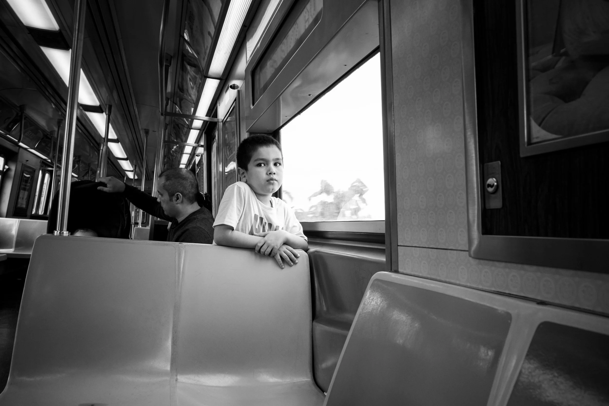 Two young boys sitting on a bus, one boy leaning on the seat, the other boy looking at the camera with a neutral expression, window showing motion blur outside.