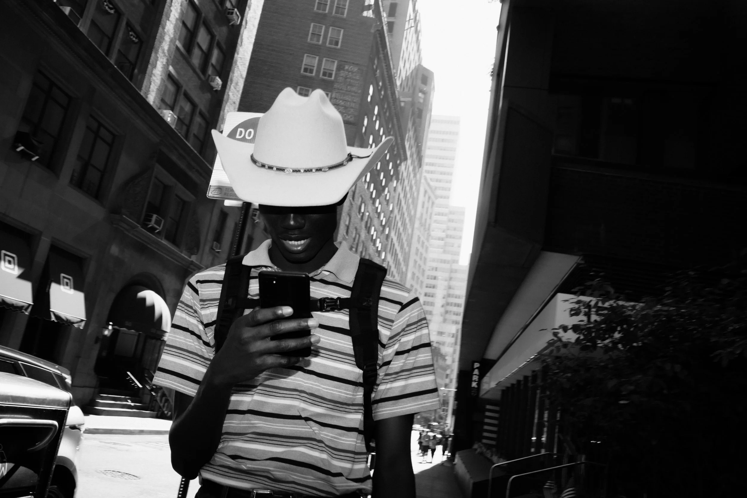 A person wearing a cowboy hat with a decorative band, striped shirt, and sunglasses, looking at their phone, on a city street with tall buildings, shops, and cars.