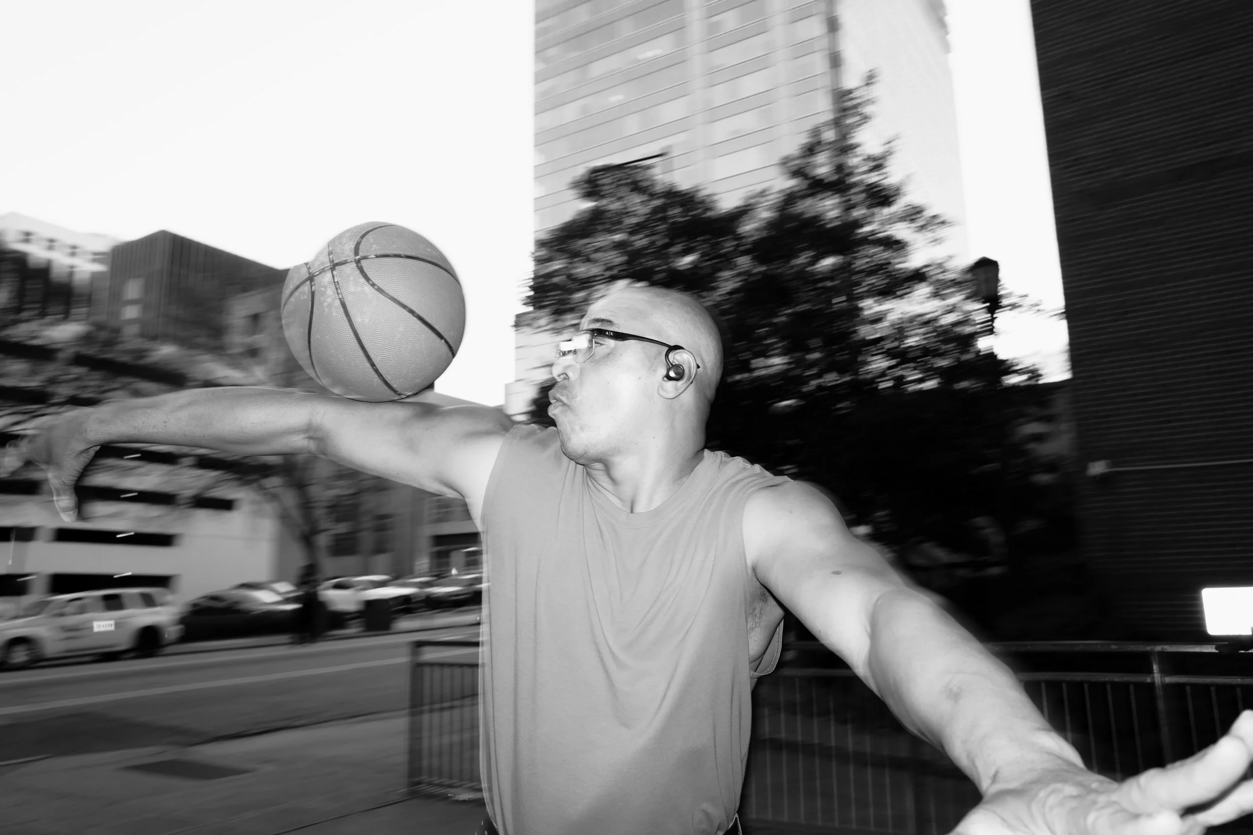 A man playing basketball outdoors in an urban setting, wearing glasses and wireless earbuds, captured in motion with a blurred cityscape background.