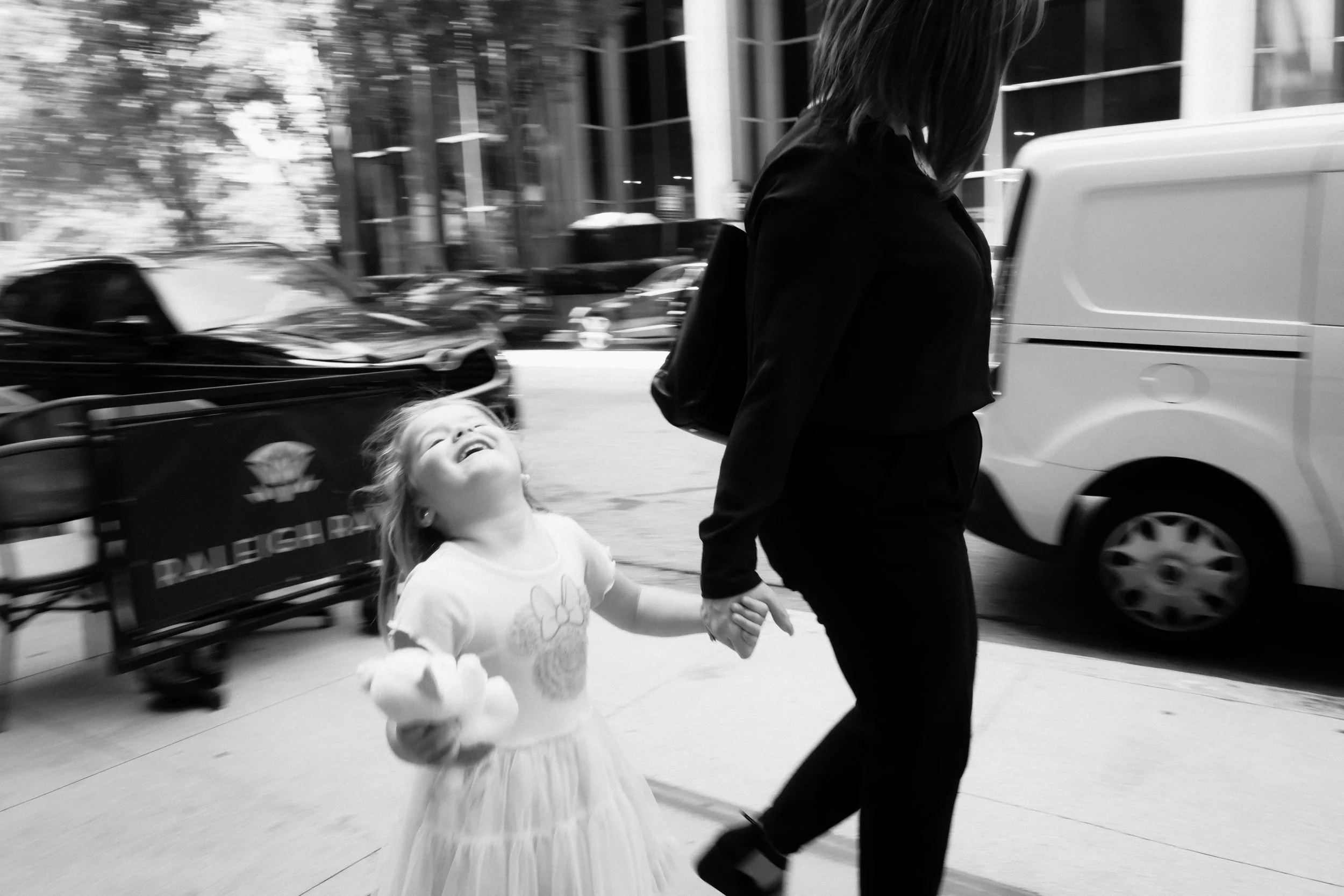 A young girl in a tutu dress smiling and looking upward while holding hands with a woman walking outside on a city sidewalk with parked cars and buildings in the background.