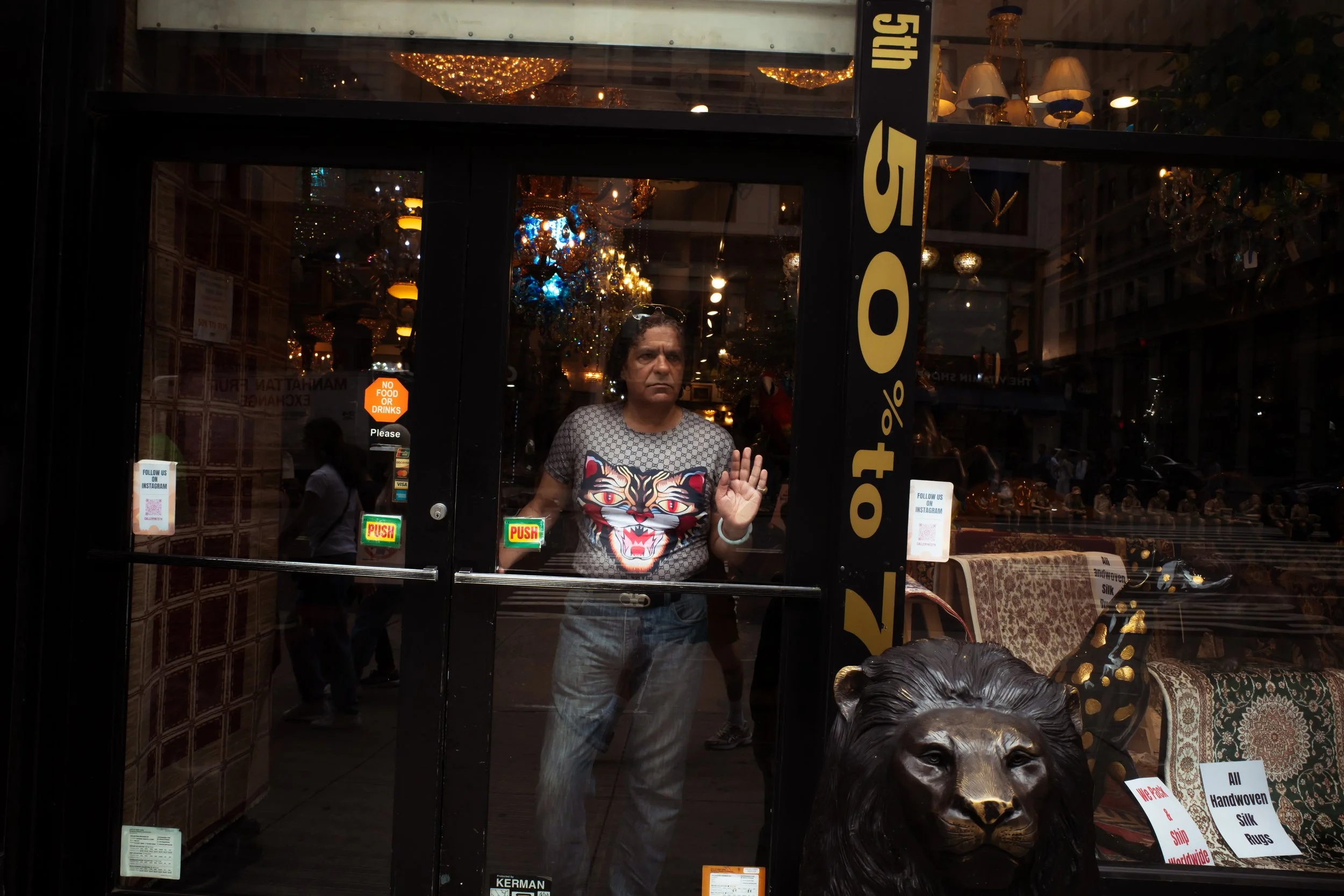 A person inside a store looking out through a glass door, with chandeliers visible inside and outside reflections, and various signs and decorations on the door.