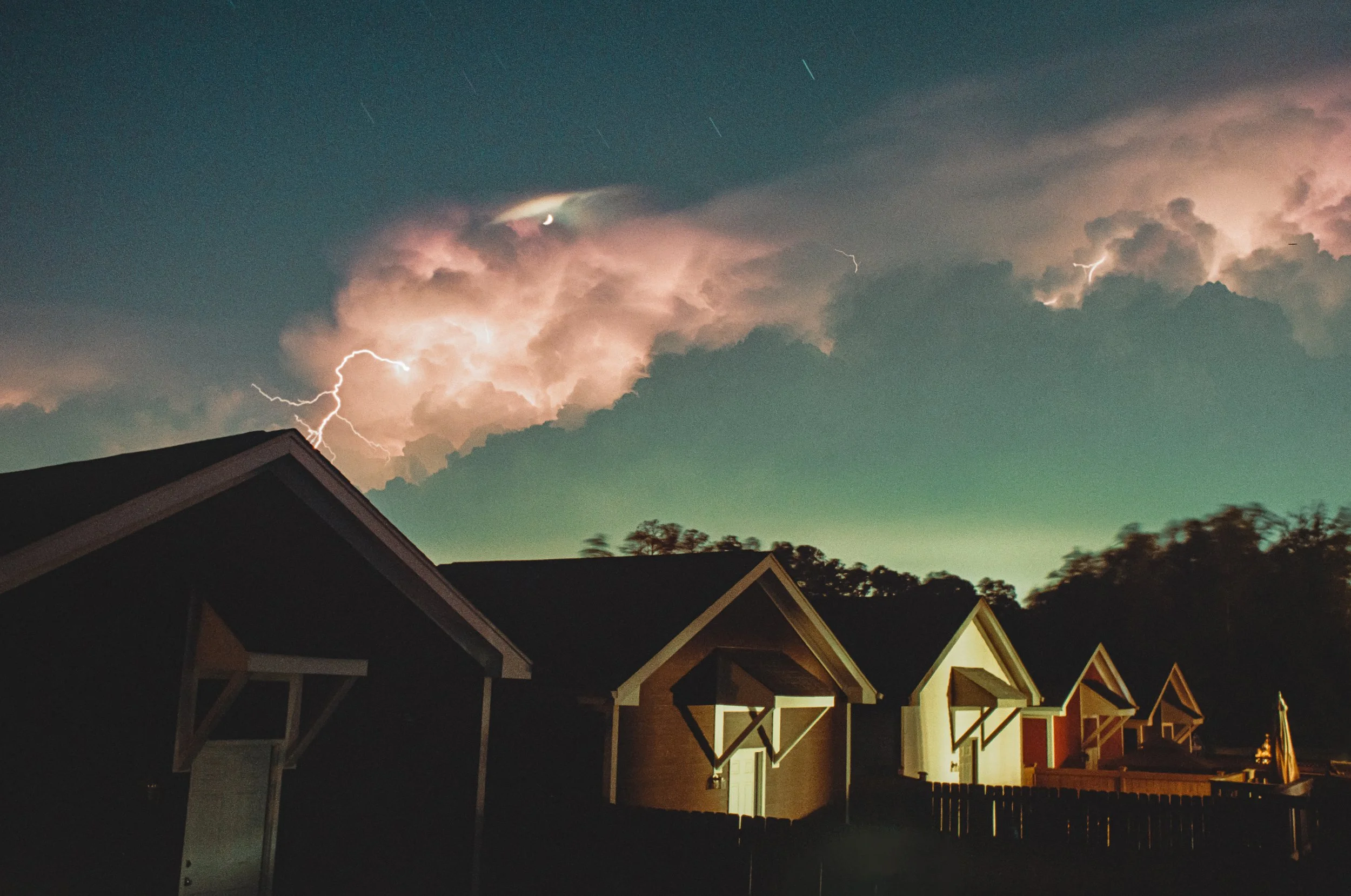 Homes with dark rooflines and a backyard, night sky with lightning, clouds, and visible stars, and a crescent moon.
